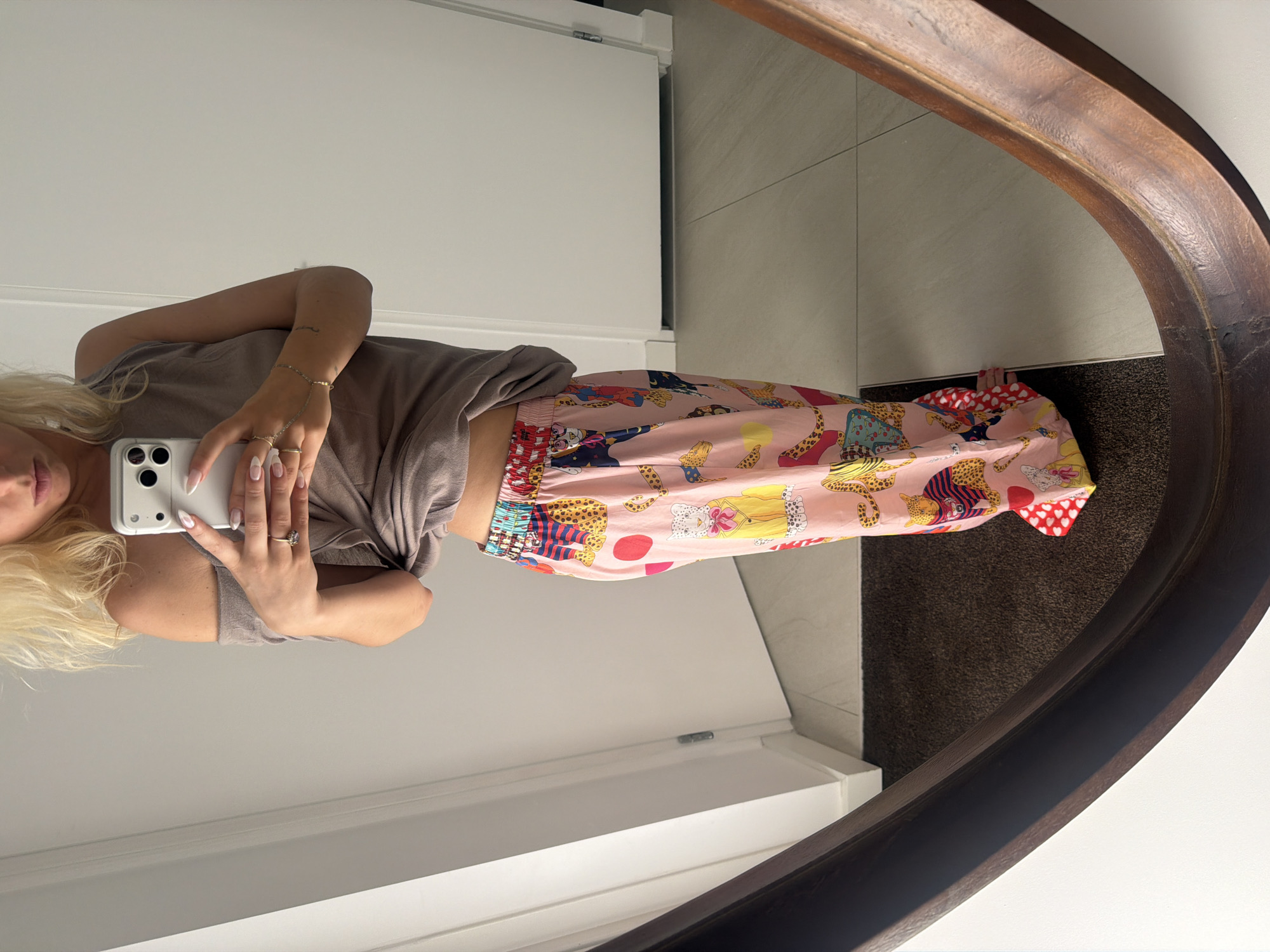 A full-length shot shows a woman standing in front of a mirror. She is wearing a taupe off-the-shoulder top and pink, patterned pants with cheetahs and polka dots. She is holding a white smartphone in her right hand, and her left hand is on her hip. The background is a plain wall with a doorway and a dark-colored rug. The image appears to be taken indoors, likely in a home, and the lighting is natural.