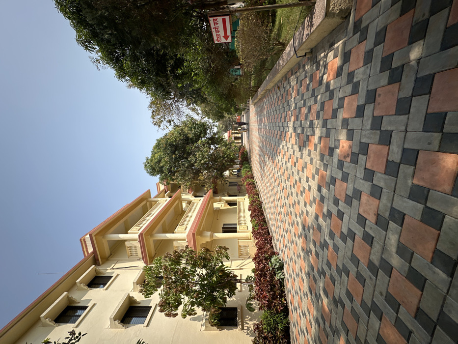 The image displays an outdoor scene featuring a patterned pathway alongside a multi-story building, set against a clear blue sky. The perspective is slightly low, looking along the pathway as it recedes into the distance.
The pathway, on the right side of the frame, is made of interlocking pavers in a distinctive geometric pattern. The pavers are primarily light grey, dark grey/black, and a warm terracotta or reddish-brown color. They form an intricate design that extends from the foreground towards the background, with direct sunlight casting sharp shadows on its surface. To the left of the pathway is a narrow strip of well-maintained shrubbery, featuring plants with both green and reddish-brown foliage.
Adjacent to the shrubbery, on the left side of the image, stands a building that appears to be at least three stories high. The building's exterior walls are a light cream or pale yellow color. It features multiple balconies with elegant reddish-brown railings and trim. Several rectangular windows with dark frames are visible on each floor. Trees with lush green foliage are interspersed along the building's side, and taller trees extend into the upper left portion of the sky.
In the mid-ground, on the right side of the pathway, a red and white sign is visible. The text on the sign, written in Hindi script, reads "सोनाली पानी मार्ग" (Sonali Pani Marg), accompanied by a white arrow pointing to the right.
The sky above is bright and clear blue, indicating sunny weather. There is no human activity evident in the scene. The overall impression is one of a well-maintained outdoor area, possibly within a park, campus, or residential complex, under bright daylight conditions.