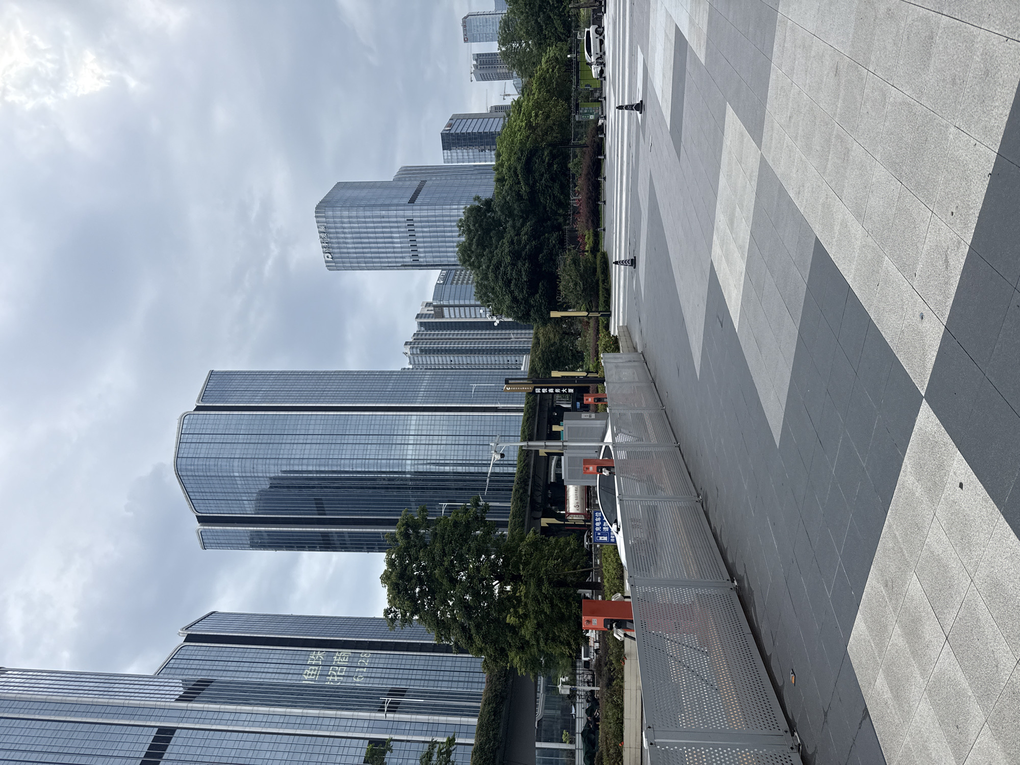 In Huangpu District, China, a modern urban landscape unfolds under a cloudy sky. The foreground is dominated by a paved plaza, with large square tiles in shades of grey and white arranged in a geometric pattern. A low, perforated metal fence runs along the left side of the plaza, separating it from a planting bed with bushes and a mature tree. Beyond the planting bed, several electric vehicle charging stations, painted bright orange, are visible.
Further into the scene, a line of contemporary glass-fronted skyscrapers rises against the overcast sky. Some of these buildings feature subtle inscriptions, such as "珠" and "招商" on one facade, along with the numbers "6128". A raised walkway or elevated transportation line is visible above the trees and behind the charging stations. In the mid-ground, a white car is parked near one of the charging stations, and other vehicles can be seen further down the road. The background reveals more buildings and hints of construction cranes, typical of a bustling city environment. The overall impression is one of a well-developed business or commercial district.