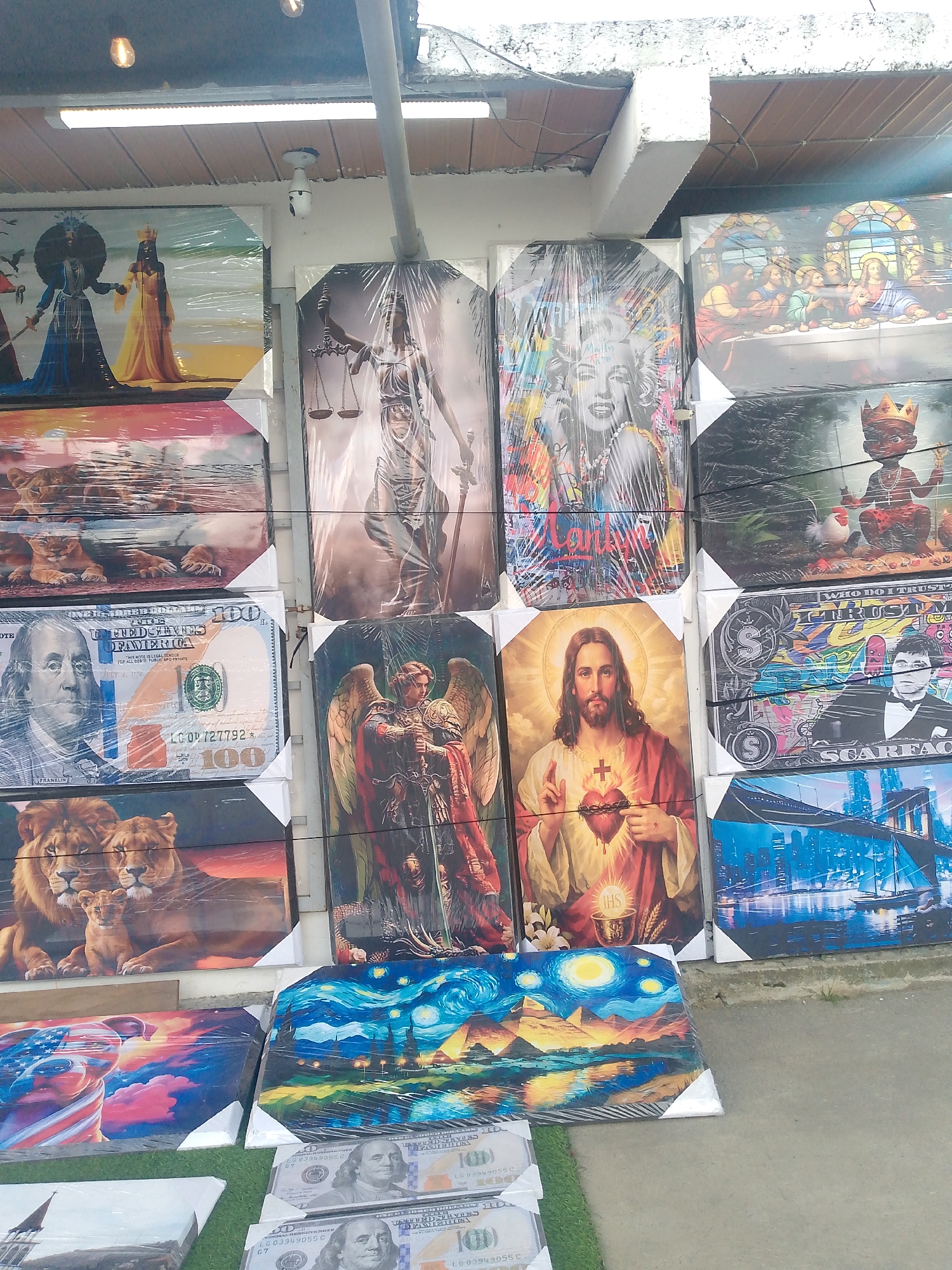The image depicts a display of framed art prints. The prints are diverse in subject matter, including religious figures like Jesus and angels, iconic figures like Marilyn Monroe, depictions of money (specifically $100 bills), animals (lions), and famous artworks such as "The Starry Night". The prints are encased in plastic wrap. The scene appears to be a street vendor's display, with some of the prints resting on the ground. Textual information within the image includes inscriptions on the money prints and the word "Scarface" on one of the artworks. The background suggests an urban environment. The photo might be taken in Caracas, Venezuela, as specified.