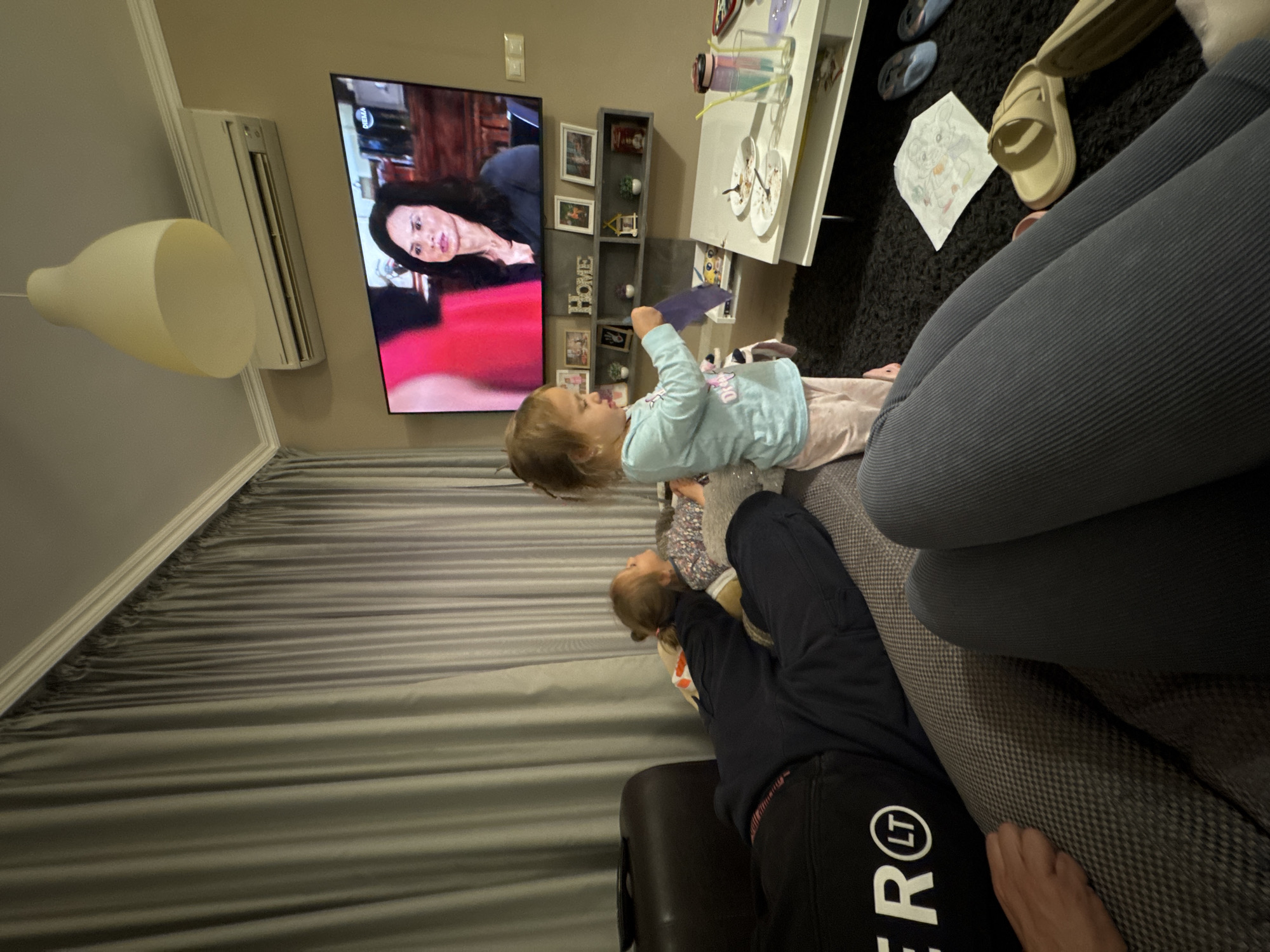The image captures an indoor scene with multiple individuals and objects, seemingly within a residential setting. A TV screen is prominently displayed on the wall, showing a woman's face. To the right of the TV is a shelf unit with photo frames and decorative items. In the foreground, a young girl in a light blue top is positioned, holding a blue object. Another child is nearby, seemingly lying on a couch. Adjacent to the children, a figure wearing gray pants and a black sweatshirt with white lettering is situated on the couch. Further to the right, there is a person's hand resting on the couch. A white coffee table with dishes is also visible, along with items like flip-flops and a drawing on the floor. The overall setting suggests a casual, domestic environment. The location is not evident based on the provided image content. The city of Varna, Bulgaria cannot be determined from the image alone.