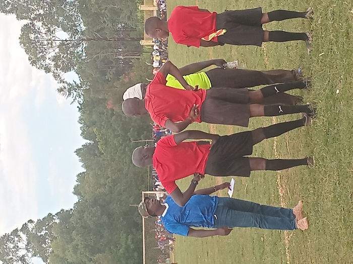 The image depicts an outdoor scene on a grassy field in Chevaywa ward, Kenya. In the foreground, five adult males are present. On the far left, a man wears a blue short-sleeved shirt, blue jeans, light brown shoes, and a camouflage print hat; he holds a white paper in his left hand and faces right. To his right, three men are dressed in matching red short-sleeved shirts, black athletic shorts, black knee-high socks, and athletic shoes, standing with their backs partially or fully to the viewer. Two of these individuals have their hands on their hips. The man on the far right has a yellow and red striped flag tucked into the waistband of his shorts. Partially visible between two of the men in red is a fifth individual wearing a lime green shirt and dark pants, holding a clear plastic bottle. In the midground, a white-netted soccer goal is visible on the left side of the field. A crowd of people is gathered in the distance across the field, partially obscured by a light-colored fence or wall on the right. The background consists of a dense line of green trees under an overcast sky.