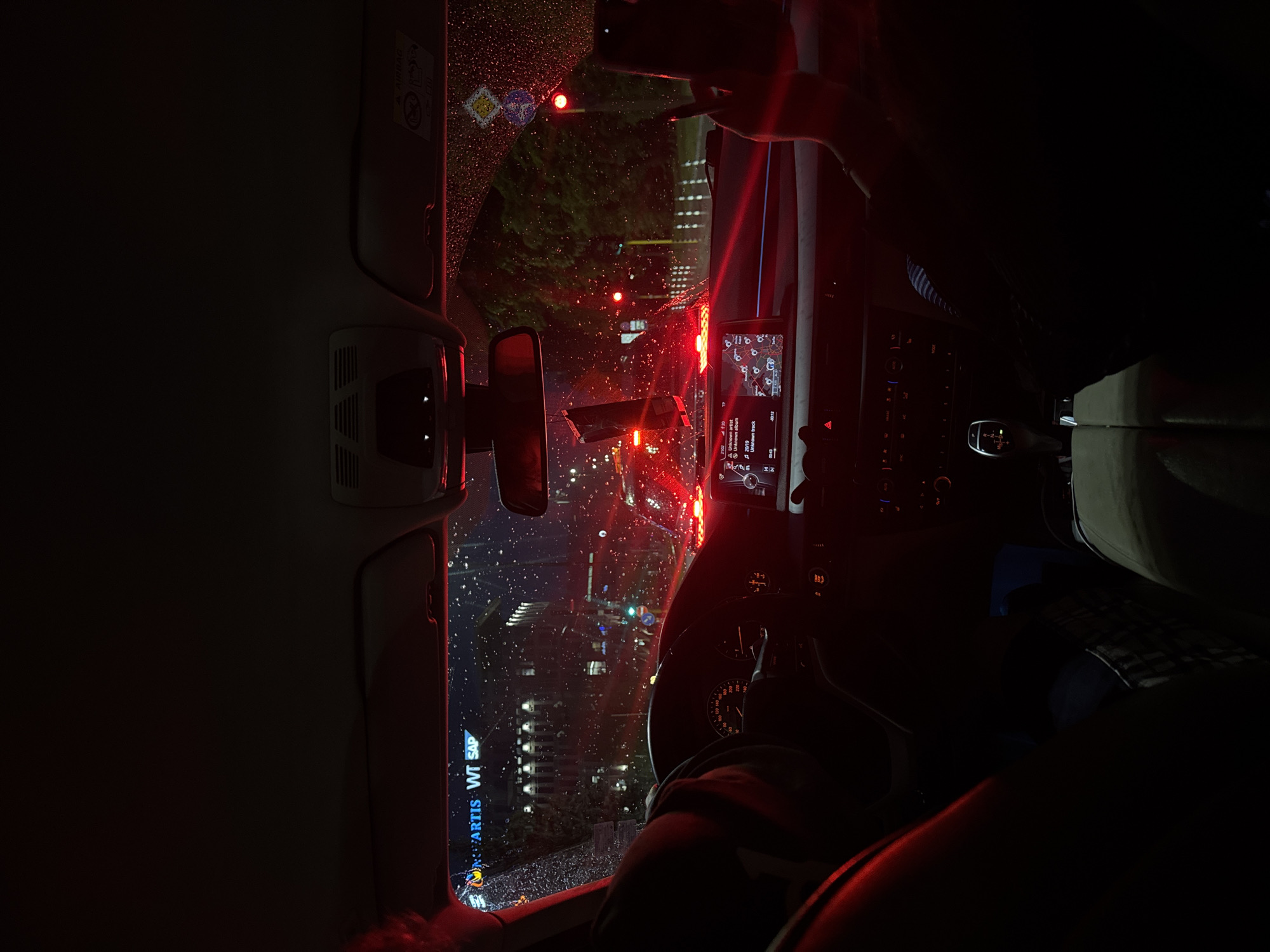 Here is a detailed description of the image:
**Setting:** The image captures the interior of a car during a rainy night. Outside the car window, a cityscape glitters with lights, blurred by raindrops on the glass. The reflection of streetlights and traffic signals is visible on the windshield. The interior of the car is dimly lit, with the red glow from the car's dashboard and ambient lighting dominating the scene.
**Subjects and Objects:**
* **Car Interior:** The dashboard, steering wheel, rearview mirror, and center console are visible. The car's infotainment system is displaying a map and music information ("Unknown artist", "Unknown album", "Unknown track").
* **Driver:** A person is seated in the driver's seat, presumably holding a phone up to take a photo.
* **Exterior View:** Through the rain-streaked windows, buildings, streetlights, traffic lights, and signage are visible. A sign for "Novartis WT SAP" can be seen through the front passenger window on the left-hand side.
**Actions and Interactions:**
* Someone in the car is taking a photo or video with their phone.
* The car is likely stopped at a traffic light, indicated by the red lights outside and the stop-and-go traffic pattern.
**Environment:**
* **Weather:** It is raining, as evident from the raindrops on the windows.
* **Lighting:** The overall lighting is low, with the red tones from the car's interior and external traffic lights creating a moody atmosphere.
**Location in Sofia, Bulgaria:**
The image was taken near a Novartis WT SAP office, which is located at Tsarigradsko shose Blvd 115, Sofia, Bulgaria.