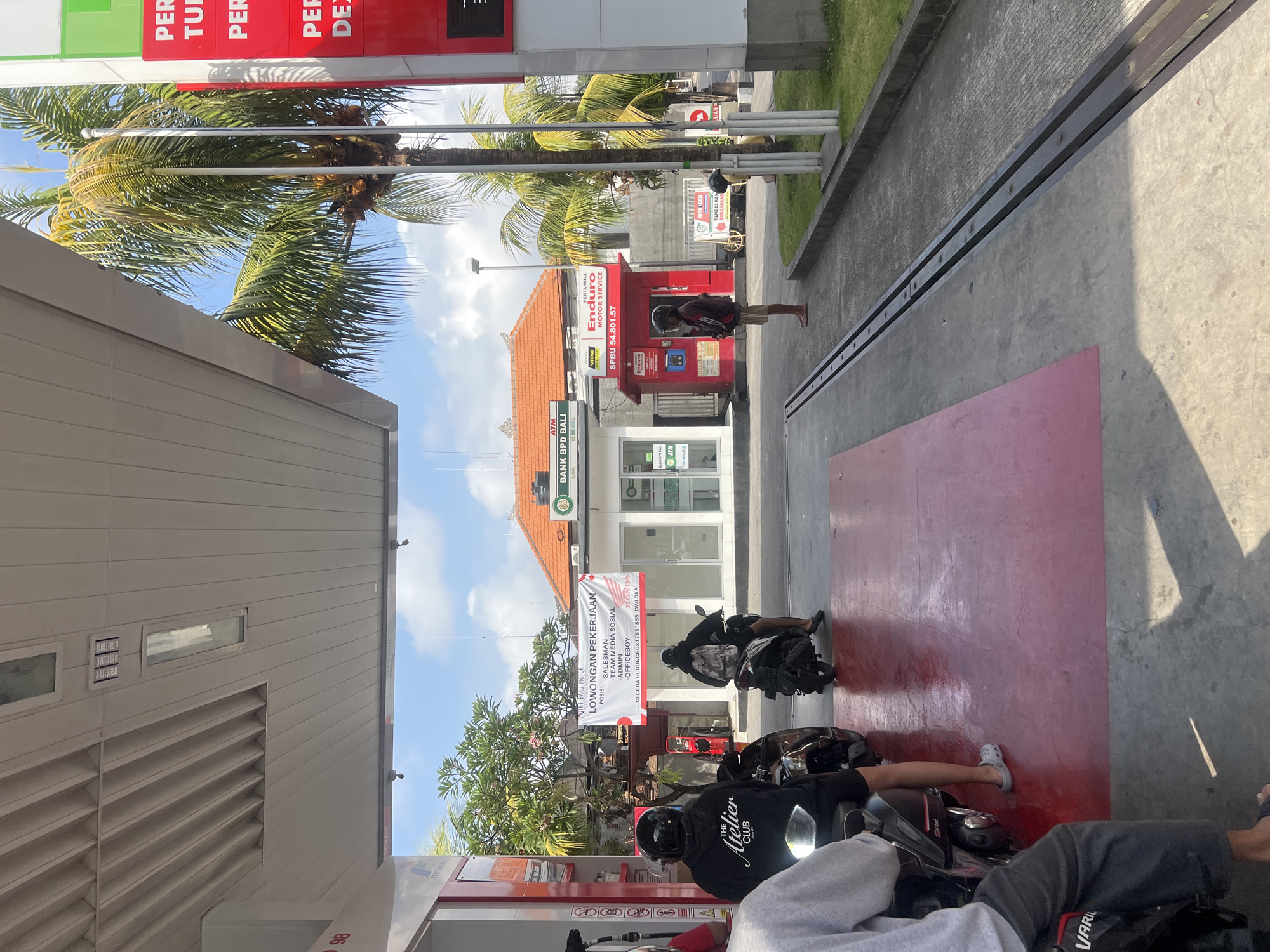 The image depicts a street scene in Denpasar, Indonesia, featuring a gas station with a prominent red and white structure. Several people are present, mostly on motorcycles. In the foreground, a person on a motorcycle is visible from the back, wearing a grey sweatshirt and dark pants. To their right, another person on a motorcycle, wearing a black helmet and a black hoodie, is also seen from behind. Further back, a man in a brown jacket and shorts stands near a red structure, and another individual on a motorcycle is partially visible in the distance.
The gas station appears to be part of a larger establishment that includes a building with the text "BANK BPD BALI" and "ATM" visible. A banner with "LOWONGAN PEKERJAAN" (Job Vacancies) is displayed, listing positions such as "SALESMAN", "TEAM MEDIA SOSIAL", "ADMIN", and "OFFICE BOY". Adjacent to this is a red kiosk with "ENDURO MOTOR SERVICE" branding and the text "SPBU 54.801.57".
Palm trees are visible in the background, suggesting a tropical climate. The ground is paved, with a red-colored section in the foreground. The sky is blue with some white clouds. The overall atmosphere is that of a typical day at a gas station in an urban environment.