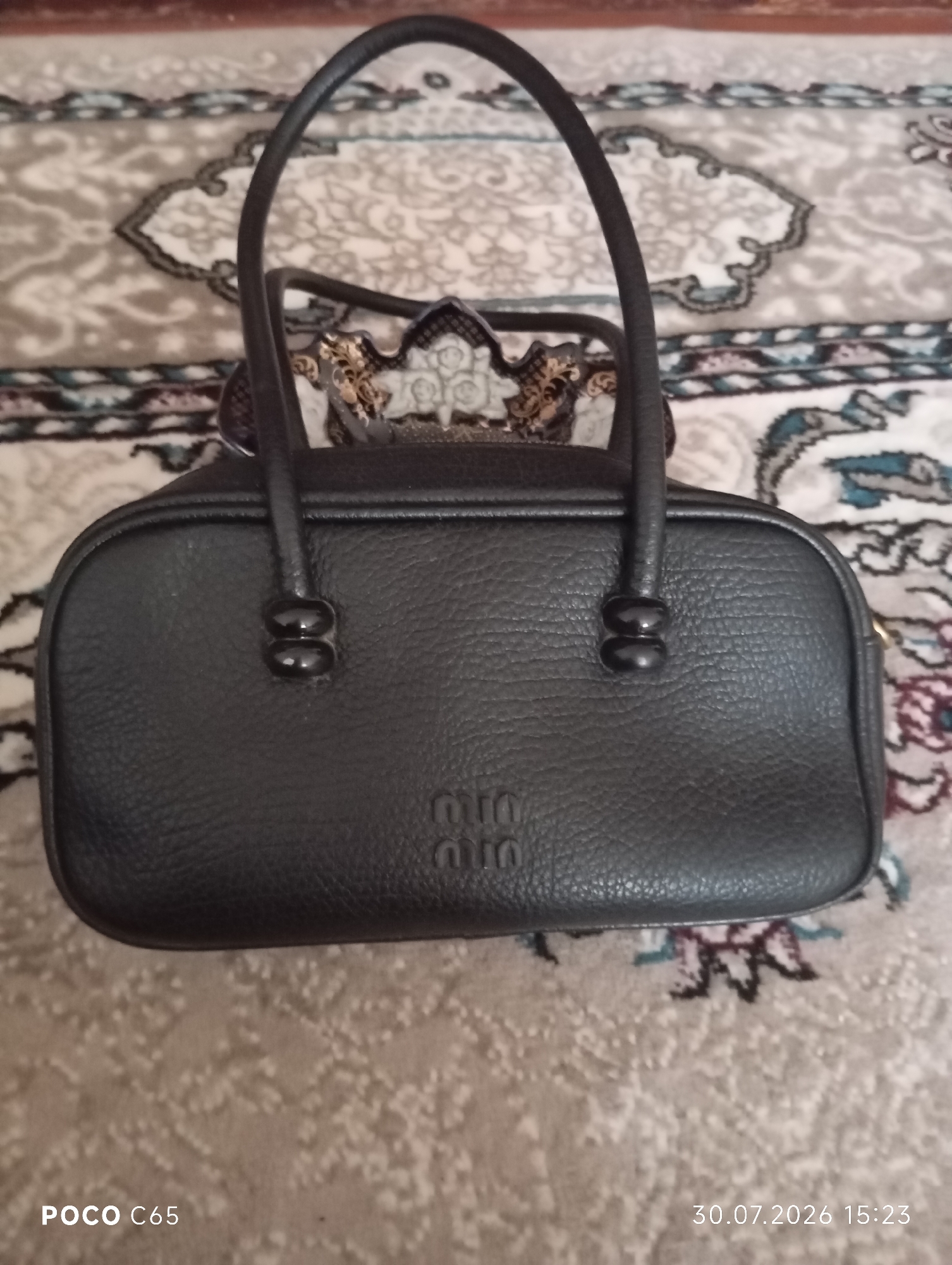 A black leather handbag with a textured finish is positioned centrally on a patterned rug. The handbag has two rounded handles and a prominent embossed logo featuring the letters "miumiu". The rug has a light beige base with intricate patterns in muted colors, including shades of gray, blue, and pink. The lighting is soft, casting subtle shadows. The timestamp "30.07.2026 15:23" is visible in the bottom right corner of the image, and "POCO C65" is in the bottom left. This image was taken in Beruniy, Uzbekistan.