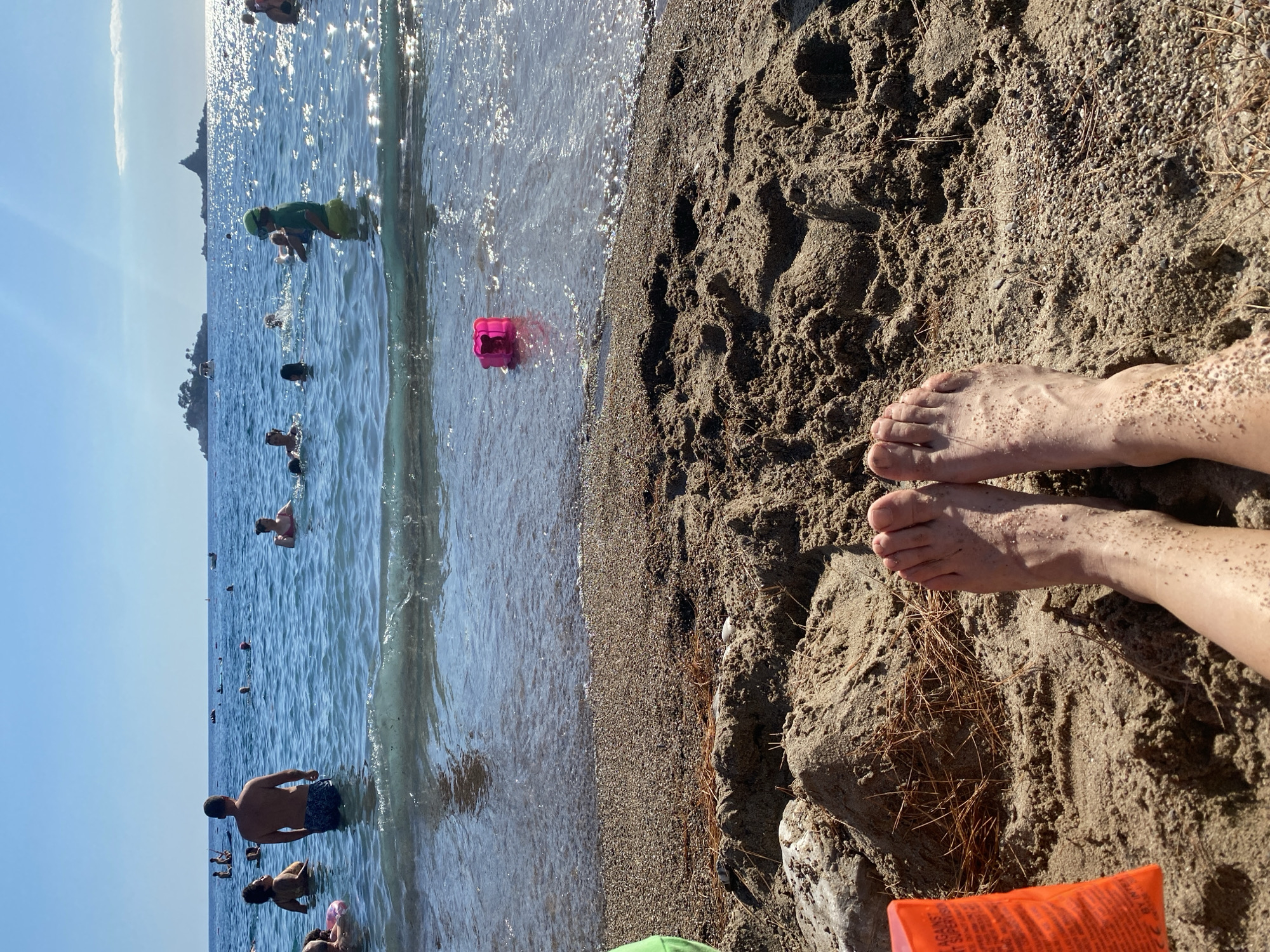 The image shows a beach scene in Petrovac centar, Montenegro. In the foreground, two bare feet are visible, covered in sand, resting on the beach. The beach is composed of sand and small pebbles, with some dry pine needles scattered on the surface.
In the middle ground, the shallow water meets the shoreline. A bright pink floating toy is visible in the water. Further out, several people are in the sea, some swimming and others standing. A man in blue patterned shorts stands in the water, facing away from the camera.
In the background, the sea extends to the horizon under a clear blue sky. Sunlight reflects brightly off the water's surface. In the distance, two small islands or rocky outcrops are visible, one with a structure on it and another with a boat near it. The overall atmosphere is one of a sunny day at the beach.