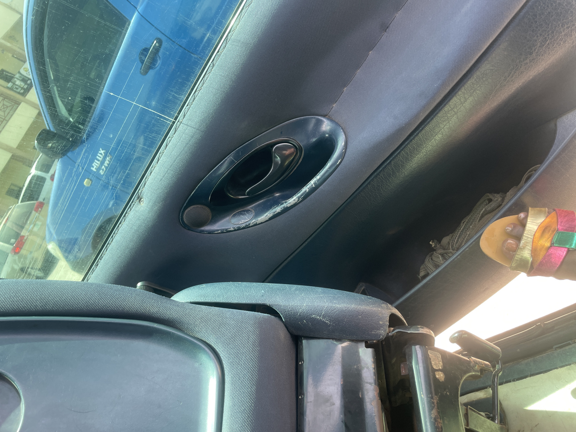 This is a close-up interior shot of a vehicle, likely a Toyota Hilux pickup truck, seen from the passenger seat looking towards the driver's side door. The dominant colors are various shades of blue and grey. The door panel is visible, featuring a black speaker grille, a chrome door handle with some wear, and a textured grey fabric insert. A portion of the grey fabric seat is also visible in the foreground, showing its textured pattern. Outside the vehicle, through the window, a bright blue truck with the "HILUX 2700" logo is partially visible. Other vehicles and buildings can be seen in the background, suggesting an urban environment, potentially a parking lot or street in Bauchi, Nigeria, given the lighting and visible architecture. A colorful sandal is also visible on the floor of the vehicle.