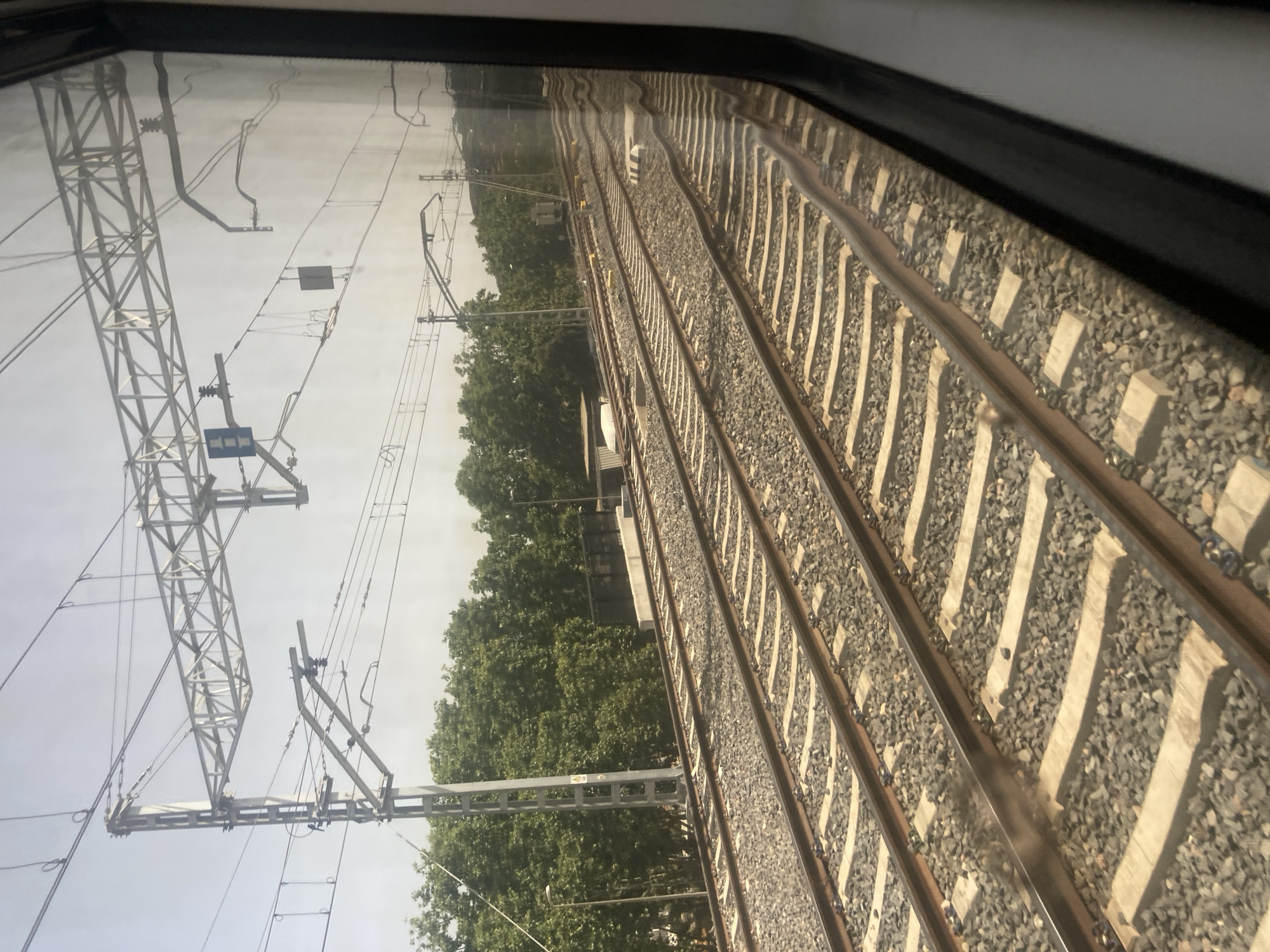 The image shows a view from a train window, looking out at multiple sets of train tracks. The tracks are ballasted with gravel and have concrete sleepers. Overhead, there are power lines supported by a metallic lattice structure and poles. Trees line the right side of the tracks, suggesting an urban or suburban environment. In the distance, a hill is visible. The sky is bright and hazy, indicating daytime. A blue traffic sign with a white symbol is visible on a pole near the overhead wires. Reflections are visible in the train window, obscuring some details. The image appears to be taken in Montcada i Reixac, Spain, a location known for its extensive railway infrastructure.