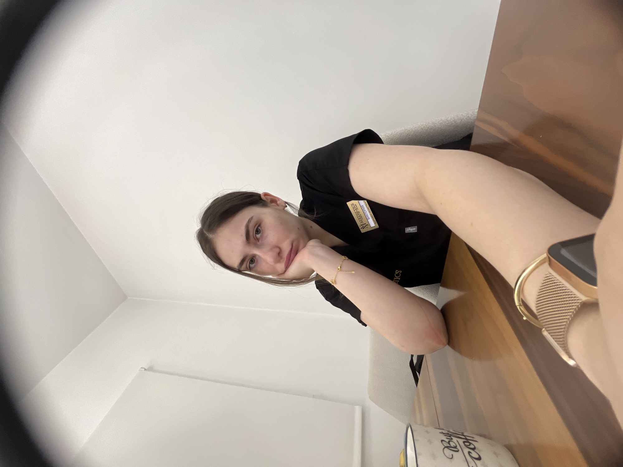 A young adult woman is captured in a close-up, angled selfie, likely taken indoors. She is seated at a glossy, dark wooden table, resting her chin on her left hand, and looking directly at the camera with a neutral to slightly pensive expression. She wears a black scrub-style short-sleeved top. A gold-toned name tag is clipped to her shirt, featuring an "N" logo, the word "AESTHETICS", and below that, "NURSE" followed by a partially obscured specialized title. The name "Saya" is visible in the bottom right corner of the name tag.
On her left wrist, she wears a delicate gold bracelet with small charms. On her right wrist, a gold-toned smart watch with a mesh band is visible. A white mug with black script text, partially reading "But first... coffee", is visible at the bottom right corner of the frame. The background consists of plain white walls, suggesting a bright, clean indoor environment, possibly an office or clinic. The lighting appears natural and bright, indicating daytime. A dark, circular element in the top left corner suggests the use of a wide-angle lens or a partial obstruction of the camera's view.