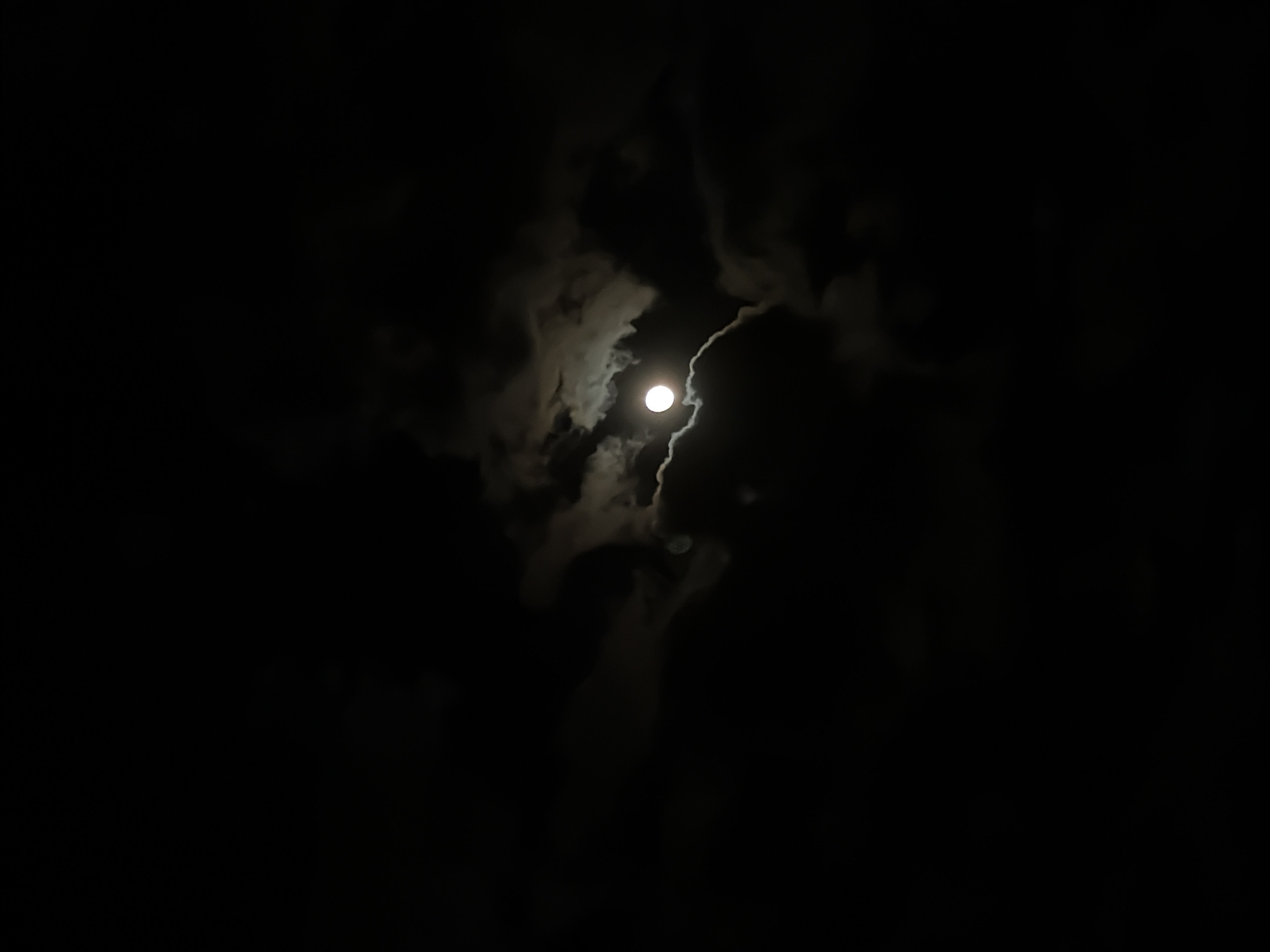 The image shows a night scene with a full moon partially obscured by dark clouds. The clouds are dense and cover most of the frame, with a brighter area where the moon is visible. The overall environment appears dark and atmospheric. There are no people or identifiable objects other than the moon and clouds visible in the frame. The location is not identifiable as Sofia, Bulgaria from the given image.