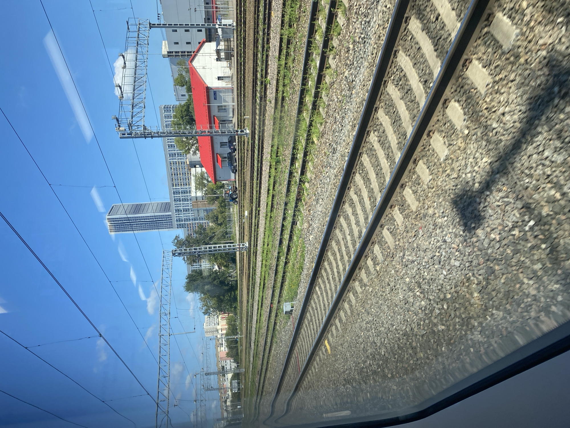 The image depicts a view from a train window, looking out at a train yard under a bright blue sky with scattered white clouds. Multiple sets of train tracks, covered in gravel and with green grass growing between them, stretch into the distance. In the middle ground, a low white building with a bright red roof is visible, along with two parked cars and a blue tractor. Further in the background, there are several multi-story buildings, including a tall, modern skyscraper. Overhead, power lines and support structures for the train tracks are prominent. The reflection of the sky and clouds is visible on the train window. The overall scene suggests an urban or industrial area with active railway infrastructure.