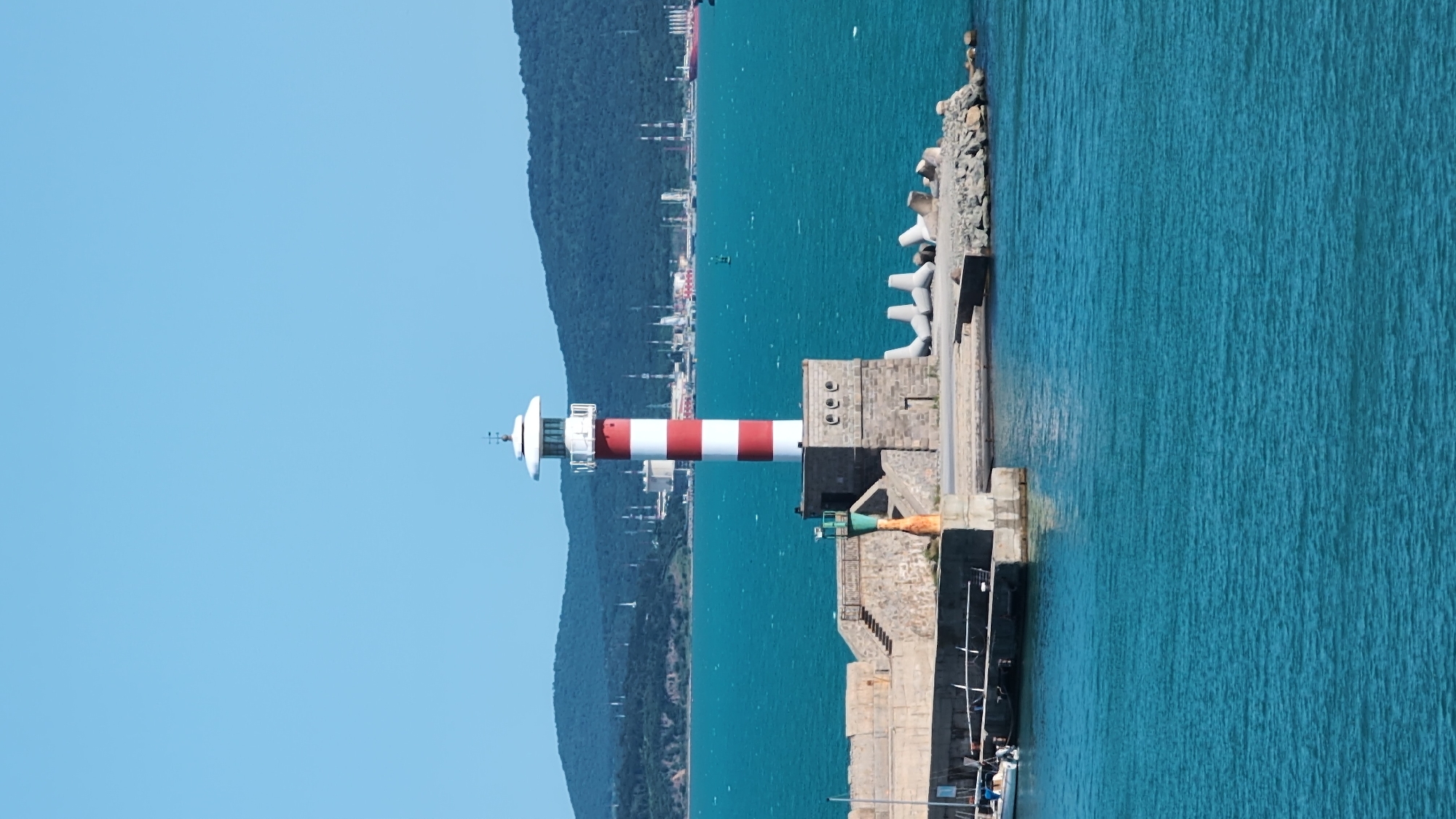The image captures a sunny day at the port of Burgas, Bulgaria, showcasing a prominent red and white striped lighthouse situated on a stone pier. The lighthouse stands tall against a clear blue sky and overlooks the tranquil turquoise waters of the Black Sea. In the background, a range of green hills with industrial structures, including tall chimneys and storage tanks, can be seen. The pier itself is constructed from rough-hewn stones, with a smaller, green-topped navigational marker located to the left of the main lighthouse. Several concrete tetrapods, designed to protect the coastline, are visible on the rocky edge of the pier. A small white boat is docked on the left side of the pier. The water is calm, with gentle ripples reflecting the bright sunlight. The overall scene depicts a functional port area with maritime infrastructure under a clear sky.