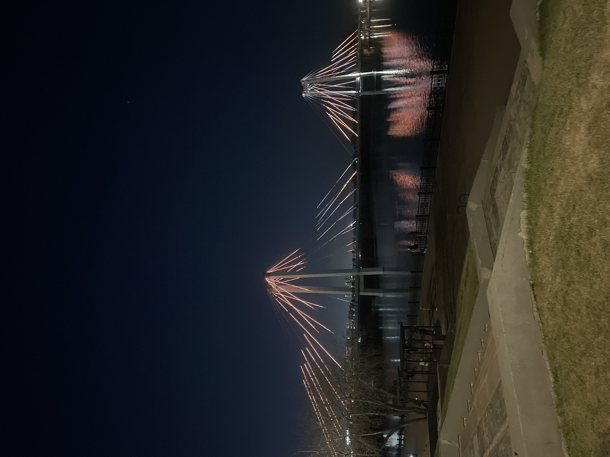 The image depicts the Vinogradovsky Bridge, a landmark in Krasnoyarsk, Russia, at night. The bridge's distinctive cable-stayed design is illuminated with lights, reflecting on the surface of the Yenisey River. On the right side of the frame, a grassy embankment and a stone walkway are visible. To the left, the sky is dark, punctuated by a single star.