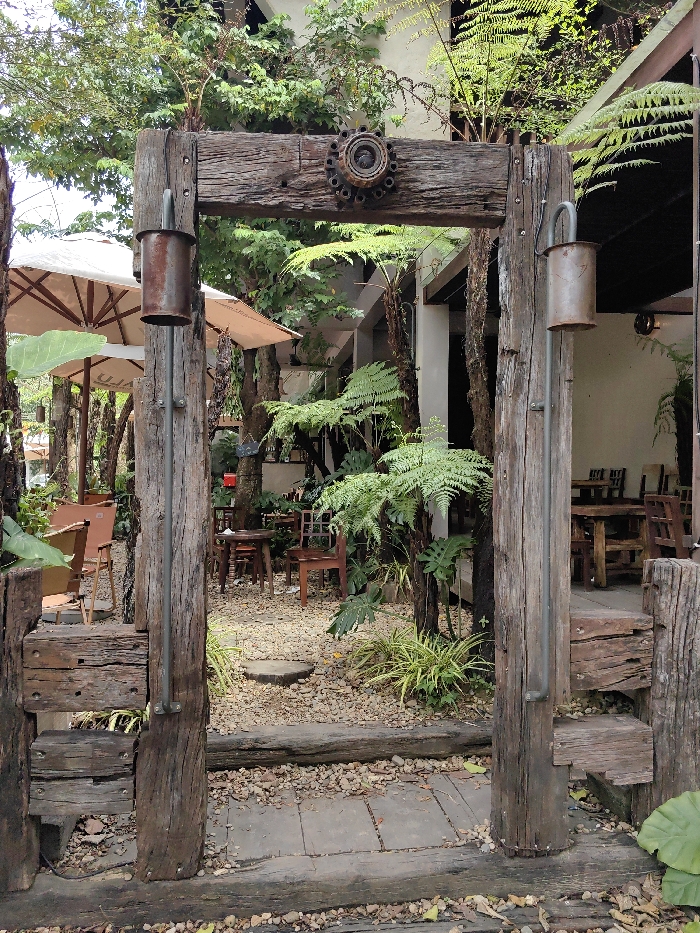 The image captures an unoccupied outdoor cafe or garden entrance located in Phú Xuân Ward, Vietnam. A prominent archway, constructed from four large, weathered dark timber beams, stands centrally. The archway features two vertical posts supporting a horizontal crossbeam, with a lower horizontal beam forming a threshold. A rusted, circular metal flange with visible bolt holes is affixed centrally to the top crossbeam. On the interior side of each vertical post, a rusted, cylindrical metal object, possibly a repurposed muffler or pipe, hangs from a curved metal rod.
Beyond the archway, a path of light-colored gravel contains a circular wooden stepping stone. To the left, several large, cream-colored patio umbrellas provide shade over multiple wooden tables and chairs. To the right, dense green foliage, including ferns and broad-leafed plants, surrounds additional wooden tables and chairs, positioned near a light-colored building with dark architectural elements partially visible. The immediate foreground shows ground covered with dark wooden planks, gravel, and scattered leaves. No individuals or active interactions are observed within the frame. The scene is illuminated by natural, diffused daylight.