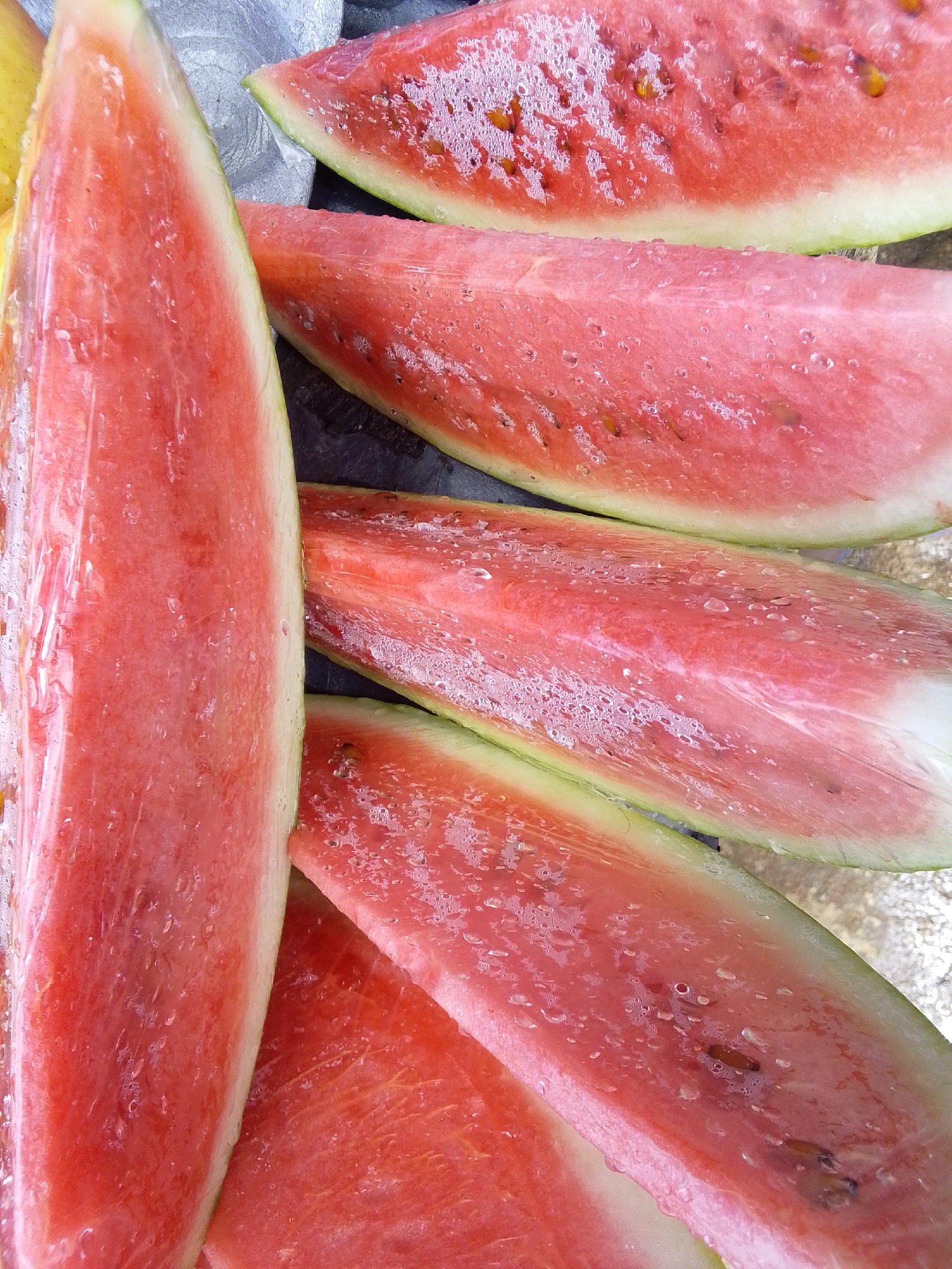 This is a close-up, overhead shot of several slices of watermelon. The slices are arranged in a staggered pattern, with the largest slice on the left filling the majority of the frame. The watermelon flesh is a vibrant pink-red, and the rind is a light green. The slices are glistening with water droplets, suggesting they have recently been cut or washed. Some of the slices show brown seeds embedded in the flesh. The background is dark and appears to be a textured surface. There is no discernible environment that would indicate a specific location in Serti, Nigeria.