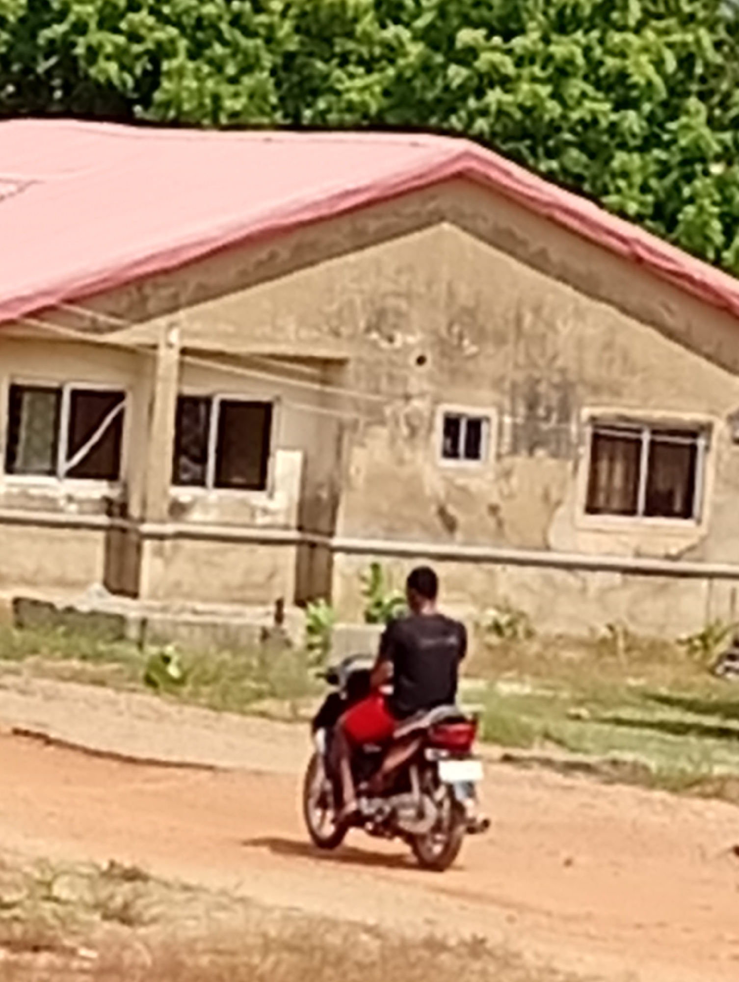 A man rides a red and black motorcycle on a dirt road in front of a one-story building with a light pink roof. The building has multiple windows, and the exterior walls appear weathered. The setting is Unknown, Unknown. Green trees are visible in the background.