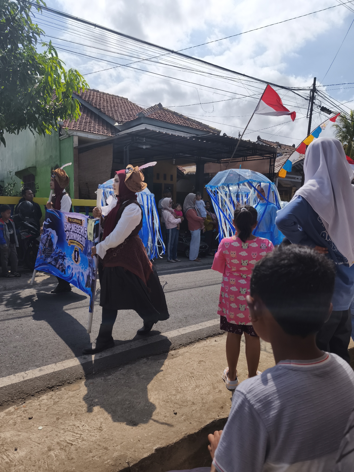 In Kutoarjo, Indonesia, a street carnival is in progress under a bright, cloudy sky. The scene captures a procession moving along an asphalt road lined with houses and trees. At the forefront, two individuals in historical costumes, including headwear with feather accents and dark skirts, hold a large banner. The banner, depicting a blue and white design with text, appears to be advertising "TIM KARNAVAL SMP NEGERI 5 PURWOREJO". Behind them, other participants are visible, including one person holding a blue umbrella adorned with white and blue streamers, resembling a jellyfish. Spectators, including children and adults, are gathered on the side of the road, observing the parade. The environment includes overhead electrical wires and flags, including an Indonesian flag, adding to the festive atmosphere.