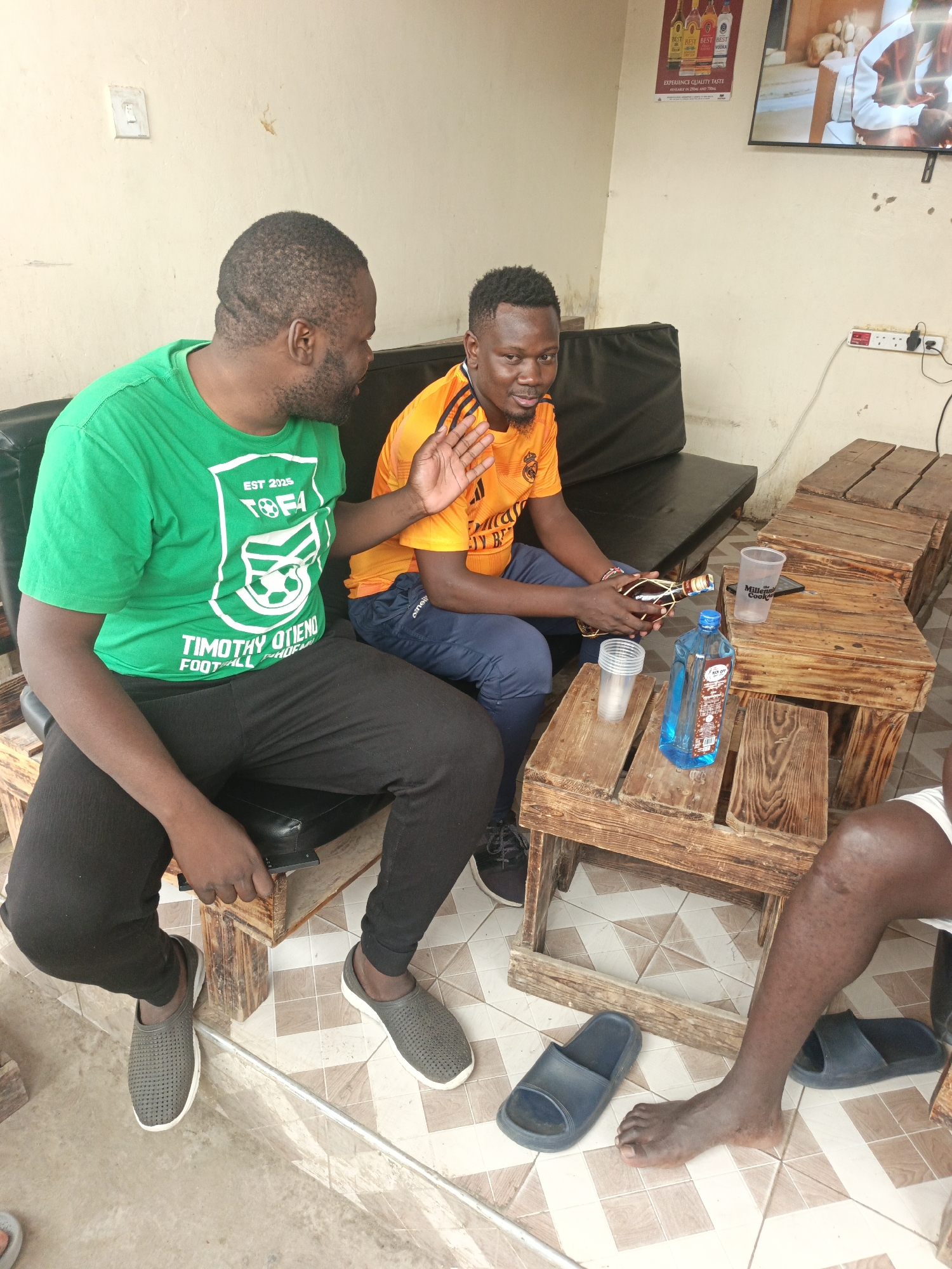 Here is a detailed description of the image:
**Scene:** The image depicts an informal outdoor or semi-outdoor social gathering. It appears to be a casual setting for drinking, perhaps in a bar or lounge area.
**People:**
* Two men are seated and engaged in conversation. One man is wearing a green t-shirt with the inscription, "TIMOTHY OTIENO FOOTBALL ACADEMY" and black pants. The other is wearing an orange t-shirt with a football logo and blue track pants.
* A third person is partially visible on the right side of the frame, only showing a bare foot and part of the leg.
**Objects:**
* There are wooden pallet tables arranged in front of the seated men.
* Alcoholic beverages are present, including a bottle of what appears to be rum and another bottle with a blue liquid (likely gin or vodka).
* Several plastic cups are on the table.
* Footwear visible includes shoes and sandals.
* A black leather sofa, which may be the sitting area for the bar.
* A television mounted on the wall in the background.
* Advertising posters for "BEST" alcohol brands.
**Actions and Interactions:**
* The two men are in the midst of a conversation. One of them has his hand raised as if emphasizing a point.
* The setting implies they are socializing and likely consuming alcoholic beverages.
**Environment:**
* The background features a cream-colored wall with a white switch or power outlet.
* The flooring is tiled in a pattern of squares, with light brown and white tiles.
* The overall setting suggests a casual and relaxed atmosphere.
**Location:**
Based on context clues in the image, such as the signage on the t-shirt, it is possible that this image was taken in Nairobi, Kenya. The man in the green shirt has "TIMOTHY OTIENO" on his shirt. Timothy Otieno is a famous soccer star from Nairobi.