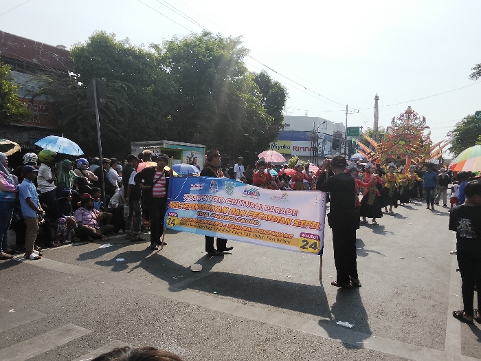 This image depicts a daytime cultural parade occurring on an asphalt street in Probolinggo, Indonesia. The scene is brightly lit, casting distinct shadows on the road surface towards the viewer.
In the immediate foreground, two individuals hold a large white banner horizontally. The banner prominently displays text in Indonesian, including "PROBOLINGGO CULTURAL PARADE" and "KOTA PROBOLINGGO," indicating the event and location. The person on the left wears a light-colored striped shirt and dark pants, while the person on the right wears a dark, formal-style suit.
To the left of these banner holders, a crowd of spectators lines the edge of the street. Individuals are standing and sitting on the ground, many holding open umbrellas of various colors (e.g., light blue, pink, red) for sun protection. Buildings with commercial signage, including partially visible "MIYAKO RINNA," are present in the background behind the spectators.
To the right of the banner, the parade procession extends down the street. A group of individuals, predominantly in traditional Indonesian dance costumes consisting of dark tops, colorful patterned skirts, and decorative headwear, are visible marching or performing. Behind them, a large, elaborate float with intricate golden and red ornamentation, possibly resembling wings or a mythical creature, is prominent. More spectators and participants, some carrying umbrellas, are visible further along the parade route.
The background features a clear, bright sky, utility poles with power lines, and dense green trees. The overall environment is an urban setting, typical of a public street event.