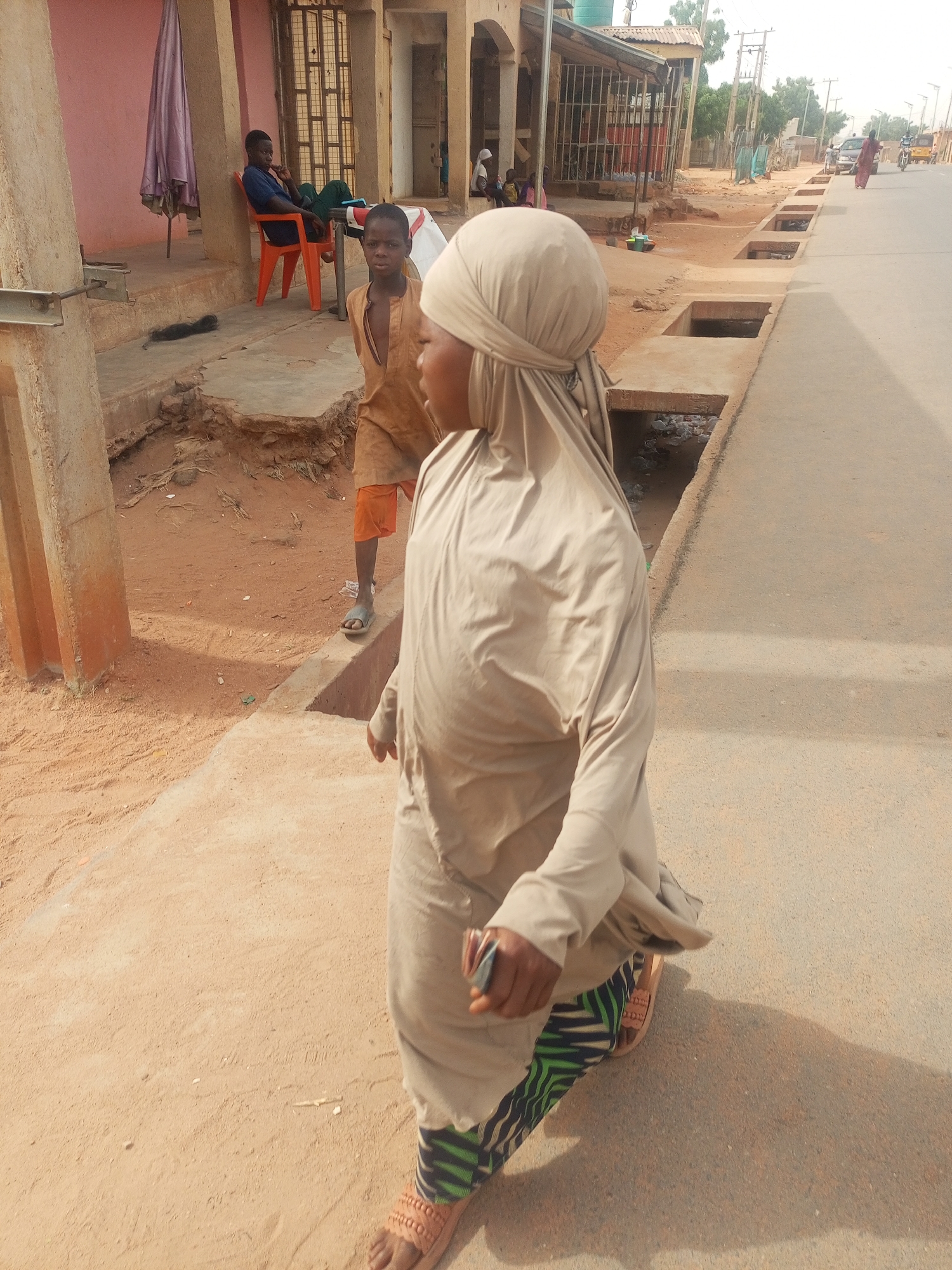 The image captures a daytime scene on a street in Tempure, Nigeria. A woman in a tan, draped garment and head covering walks in the foreground along a dirt and concrete path beside a paved street. Her sandals are visible, revealing patterned leggings. In the background, a young boy wearing a tan shirt and orange shorts walks in the same direction on a dirt path. A man is seated in an orange plastic chair in front of a pink building on the left. In the distance, there are buildings with metal doors, concrete drainage channels along the street, and other figures and vehicles farther down the road. The environment appears dusty with sandy colored soil.