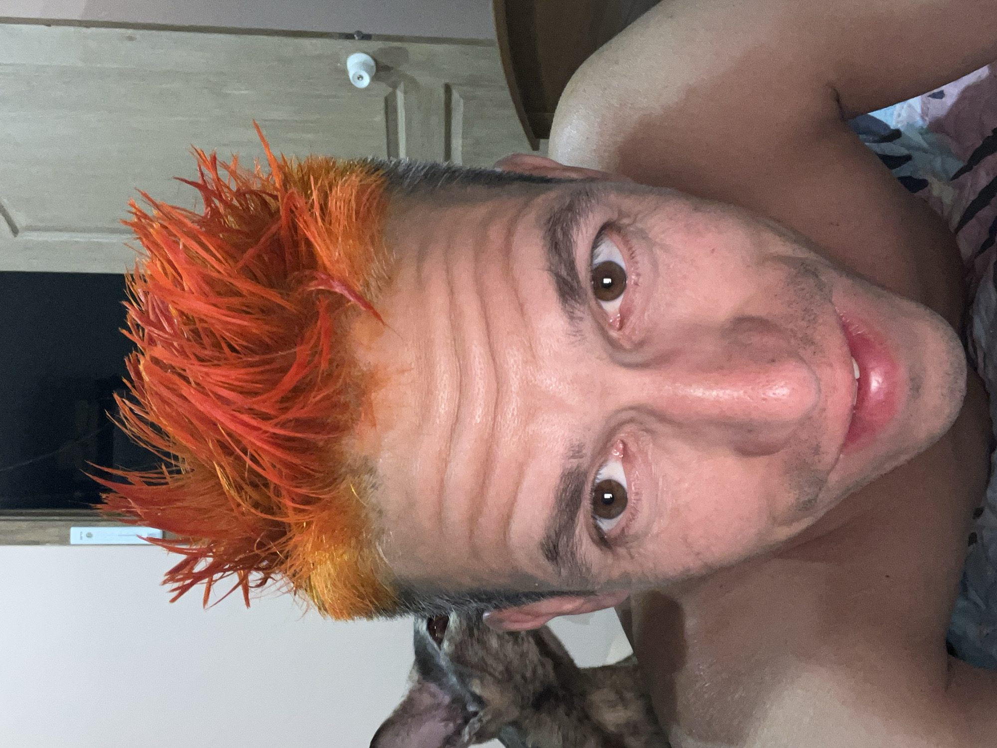 A close-up, slightly angled shot captures a man with bright orange, spiky hair. His facial features are prominent, with wrinkles on his forehead and light brown eyes. A dog is partially visible near his ear. In the background, a white door with a security camera is visible, along with part of a wooden headboard and a patterned bedspread. The man's skin appears moist. The location is unknown. The shot does not identify that the image was taken in Chaikovsky, Russia.