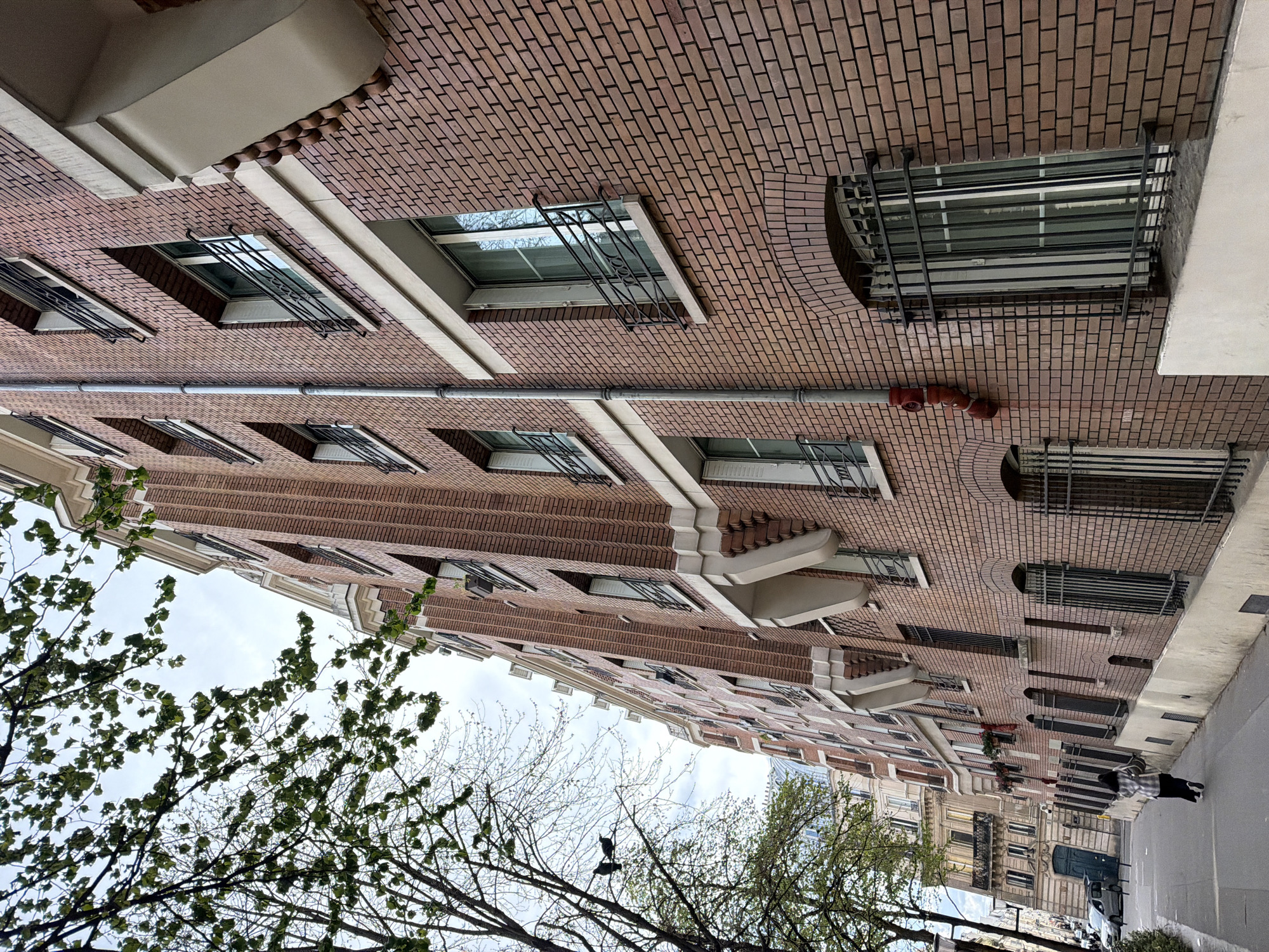 An eye-level photograph looking up at the facade of a brick building, with portions of bare tree branches visible in the foreground, likely taken in Paris, France. The building features repeating rectangular windows with metal security bars, interspersed with rounded window openings with similar bars. A horizontal pipe runs along the brickwork, and architectural details such as ledges and rounded brickwork are apparent. In the distance, other buildings and a street scene are visible, with people and cars present. The sky is overcast, and the overall tone is urban and mundane.