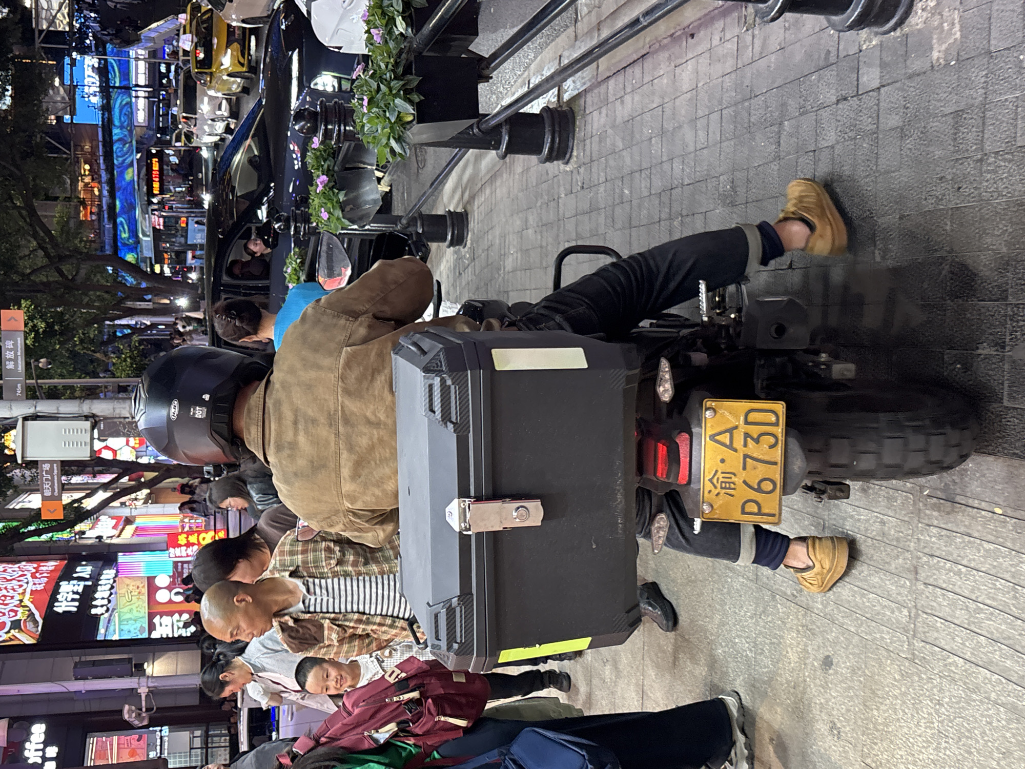 The image shows the back of a motorcycle parked on a street, with the license plate "渝・A P673D" visible. The rider is wearing a helmet and a brown jacket. A black storage box is attached to the back of the motorcycle. Several pedestrians are nearby, including a man in a striped shirt and a child. Neon signs in Chinese characters can be seen in the background. Signs in the background are labeled “朝天门广场Chastainness Square“ and “解放碑 Liberation Monument.” The location is in the Yuzhong District of China.