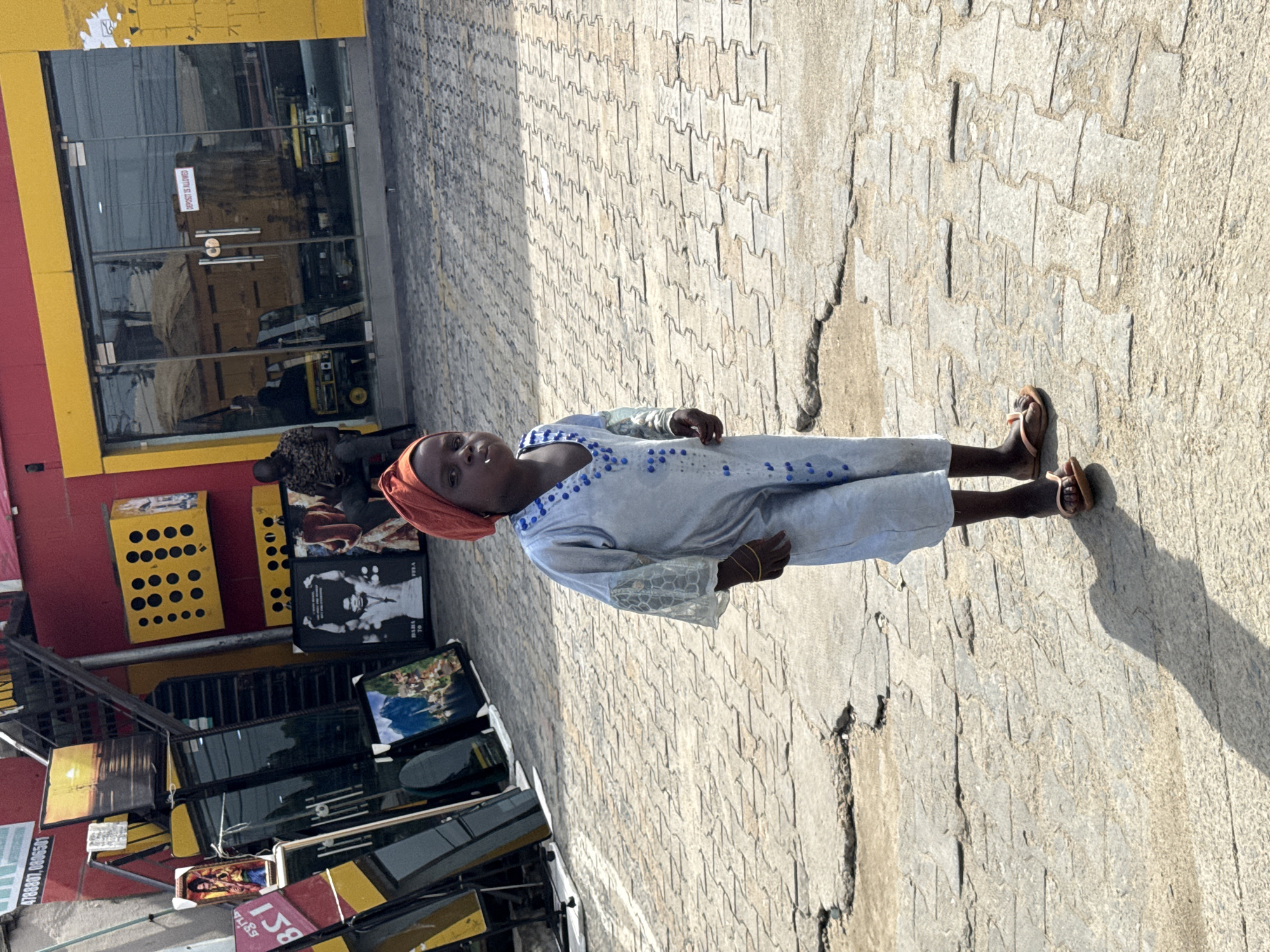 A person wearing a light-colored, long-sleeved garment with blue and white trim and a rust-colored head covering is standing on a paved area. The pavement is composed of interlocking concrete pavers and has a crack running through it. The person is wearing brown sandals. Behind the person is a building with a red and yellow facade. A shopfront with glass windows is visible, reflecting the surroundings. In front of the building, there is a display of what appears to be framed pictures or artwork. There are also some metal frames stacked nearby. A sign is partially visible in the lower left corner. The location appears to be an urban setting, possibly in Lagos, Nigeria, based on the clothing style and architectural details.