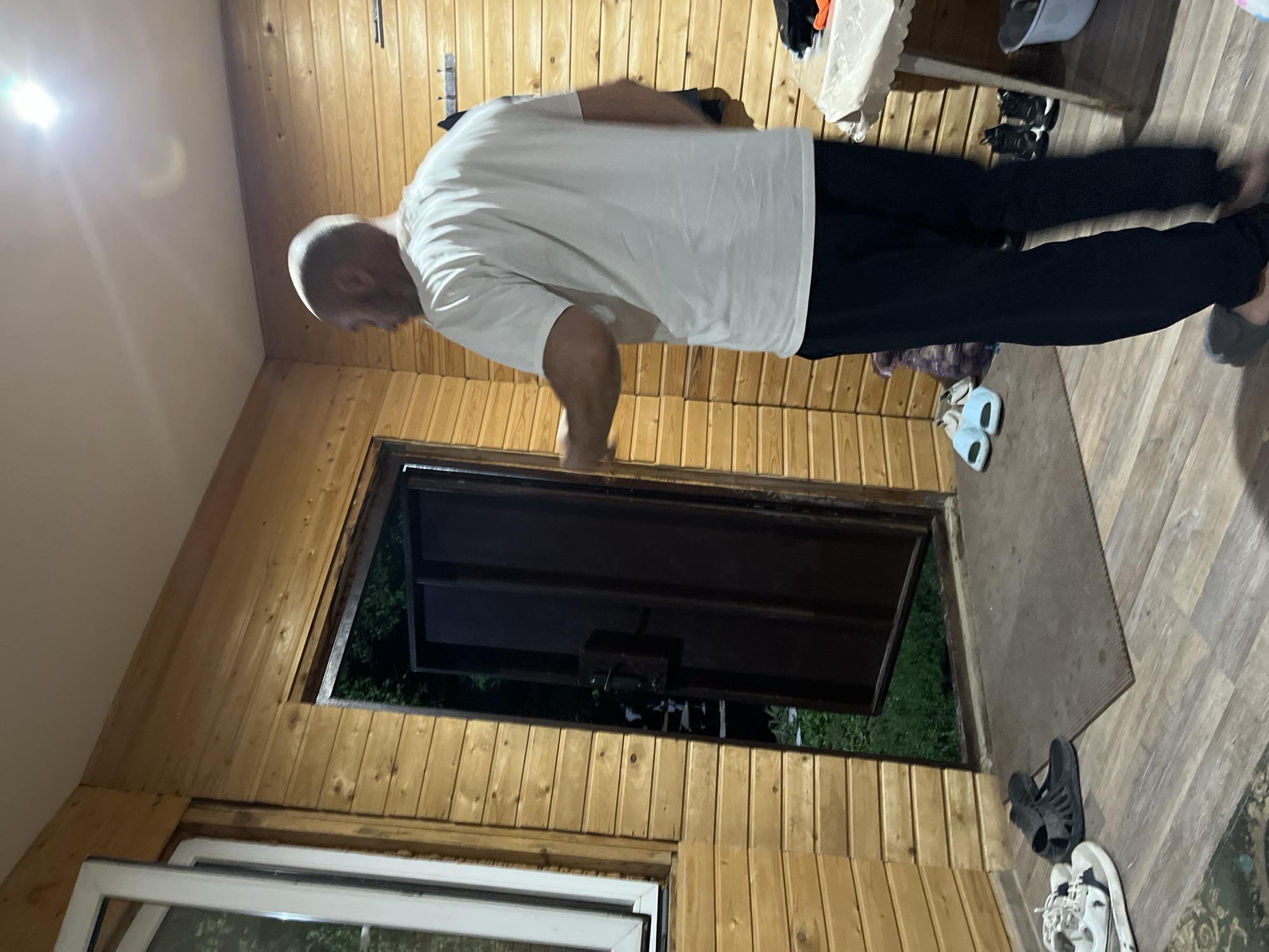 The image depicts an interior scene of a wooden structure, possibly a porch or a room with wooden paneled walls. A bald man wearing a white t-shirt and dark pants stands with his back to the camera, facing an open doorway. The doorway reveals an outdoor scene with greenery. On the floor near the doorway, there are several pairs of footwear, including sandals and sneakers. To the right, a table covered with a white cloth is visible, with some items on it, including a bucket and what appear to be shoes. The lighting suggests it is either evening or the structure is dimly lit, with a bright overhead light illuminating the ceiling. The style of the wooden construction and the types of footwear suggest a rustic or informal setting, potentially in a dacha or a similar residential outbuilding. There is no specific visual information in the image that would allow for the identification of the city as Saratov, Russia.