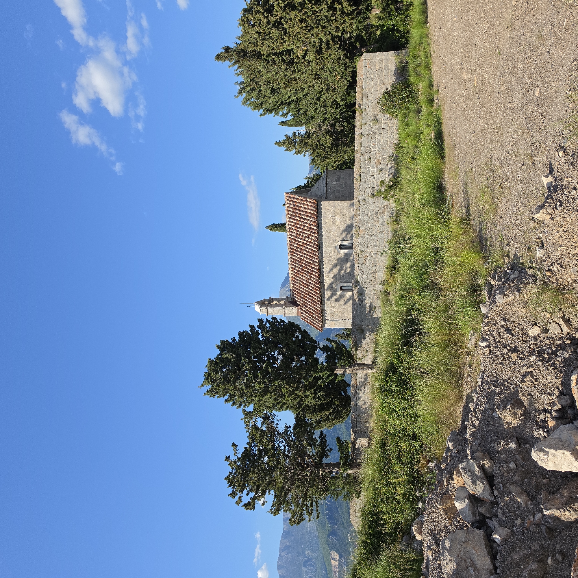 This image captures a serene landscape in Luštica, Montenegro, on a bright, sunny day, likely morning or late afternoon given the clear light. The main subject is a small, old stone building, possibly a church or chapel, with a distinctive terracotta tiled roof and a modest bell tower topped with a cross. Two arched windows are visible on its side.
The building is nestled among tall, dark green coniferous trees, which appear to be cypresses, providing a verdant contrast to the stone structure. A prominent tree stands to the left of the building, and others are visible behind it and along a stone wall that runs alongside. In the background, rolling mountains rise under a clear blue sky, which features scattered white cumulus clouds.
In the foreground and extending along the right side of the frame, there is a rustic dirt and gravel path or road. Lush green grass and various shrubs grow beside this path and near the stone wall. Loose rocks and earth are visible on the ground, adding to the natural, untamed feel of the environment. The scene is tranquil and evokes a sense of history and natural beauty. No discernible text is visible in the image.