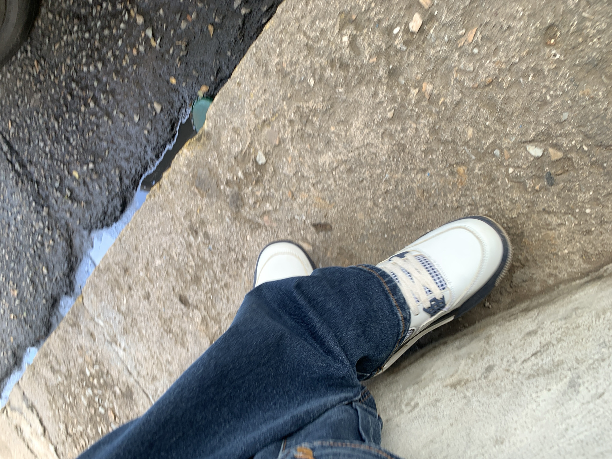 A low-angle, first-person perspective shot captures a person's legs and feet standing on a concrete surface, likely a sidewalk or curb, in Santa Ana, Venezuela. The person is wearing dark blue denim jeans and white sneakers with dark blue accents and a light-colored checkered pattern on the sides of the lacing. The left foot is slightly obscured by the right leg.
To the left of the concrete, there is a darker, textured ground, possibly asphalt or wet soil, with a visible puddle or standing water reflecting some light. A small, dark green object, possibly a plastic cap or part of a bottle, is partially visible at the edge of the puddle near the concrete. The concrete surface appears worn and has small pebbles or debris scattered across it. The lighting suggests it is daytime. No text is visible in the image.