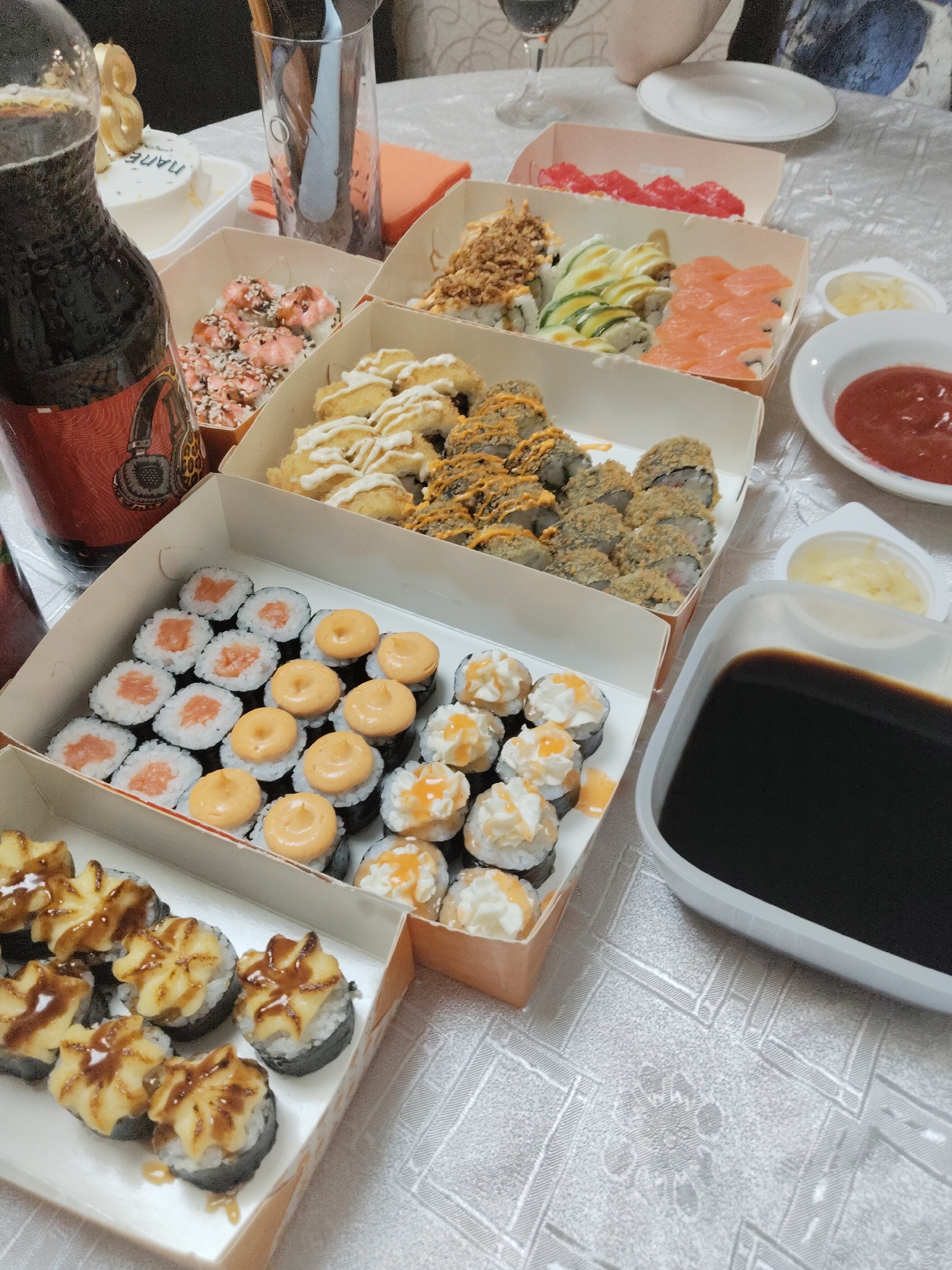 An overhead shot captures a table laden with an array of sushi, sauces, and drinks, suggesting a casual dining event, possibly a birthday celebration. The table is covered with a white, intricately patterned tablecloth.
The central focus is on numerous types of sushi rolls presented in cardboard takeout boxes. Varieties include classic maki rolls with salmon, rolls topped with creamy white and orange sauces, fried or tempura-style rolls, rolls with green cucumber or avocado slices and white drizzle, and rolls featuring raw salmon pieces. Another box holds rolls with a pink or red topping and sesame seeds, while others display rolls with a star-shaped yellowish topping and brown drizzle.
Accompanying the sushi are several condiments: a dark brown liquid in a rectangular container (soy sauce), a small white dish with red sauce (likely ketchup or sweet chili), and another small white dish containing yellow pickled ginger.
A dark-colored beverage bottle with a red label featuring a headphone graphic stands prominently on the left. A clear glass, holding chopsticks, is visible behind the bottle. To the far left, a small white cake is partially visible, topped with a golden candle shaped like the number "3". The word "ПАПА" (PAPA) is discernible in light blue icing on the cake. A white plate is seen on the right, along with a partial view of a person's arm or torso, indicating human presence. The scene is brightly lit, likely indoors.