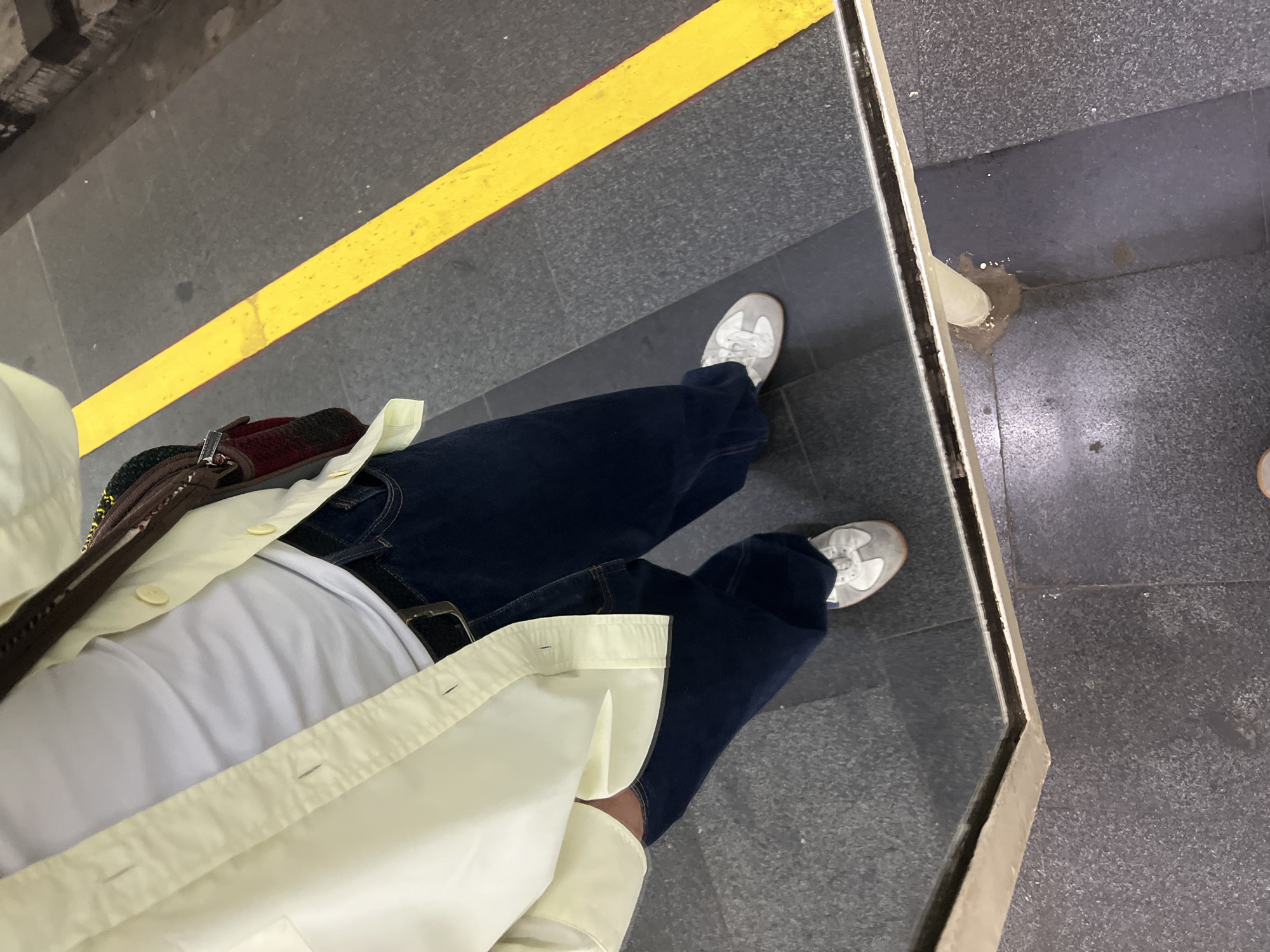 A first-person perspective image of a person standing on a subway platform. The platform floor is composed of gray tiles with a yellow safety stripe painted near the edge. The person is wearing blue jeans, white sneakers, a white t-shirt, and a light yellow button-down shirt. A bag with a brown strap is slung over the person's shoulder. The platform edge is visible on the right side of the image. A small amount of spilled liquid is near the platform edge. The location is an underground subway station platform. There is no evidence in the image to indicate it is in Yerevan, Armenia.