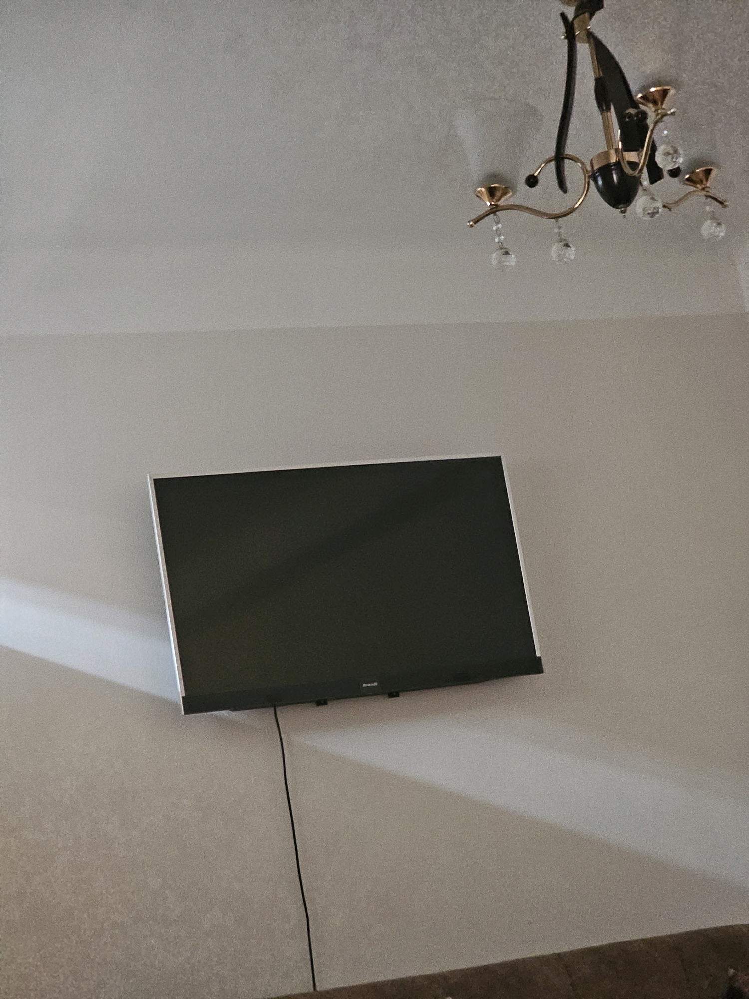 The image depicts an interior scene, likely a living room, with a flat-screen television mounted on a textured, light-colored wall. Above the television and slightly to the right, a chandelier with multiple arms and crystal embellishments hangs from the ceiling. The room appears to be sparsely furnished, with a portion of a brown sofa visible at the bottom of the frame. There are no people present in the image. The lighting suggests ambient room light, with shadows cast on the wall by the television and the chandelier. The image does not provide any specific clues to pinpoint its location in Batna, Algeria.