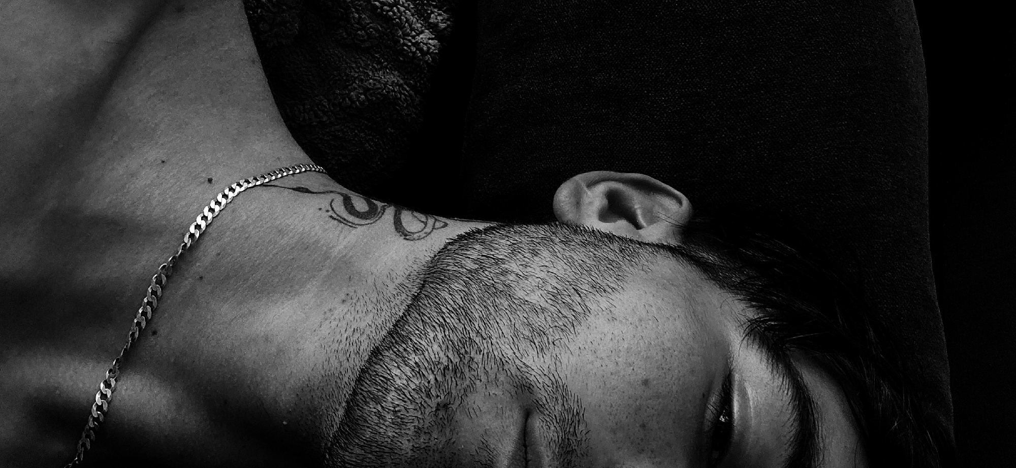 A black and white close-up image shows a person laying down, with their head slightly tilted back against a dark, possibly cushioned, surface. The person has dark hair and a beard. The right ear is visible, pressed against the dark surface. A silver chain is draped across the person's chest and neck. There is a tattoo visible on the upper neck, near the shoulder. The eye is open, but partially obscured by the upper eyelid. The image does not contain any information that is location specific.