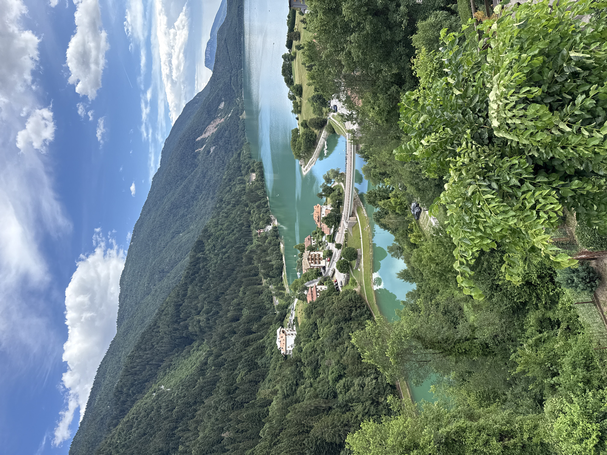 An elevated, wide-angle shot captures a picturesque scene of a bright, clear day with a scenic lake and lush green mountains in Molveno, Italy. The lake's turquoise water is tranquil and reflects the sky. On the shore, small buildings with red-tiled roofs are nestled amongst the trees. A bridge spans a narrow inlet of the lake, connecting the two sides. The foreground is dominated by dense green foliage, with a fence visible at the bottom. The sky is a vibrant blue with scattered white clouds. The overall impression is one of natural beauty and peacefulness.