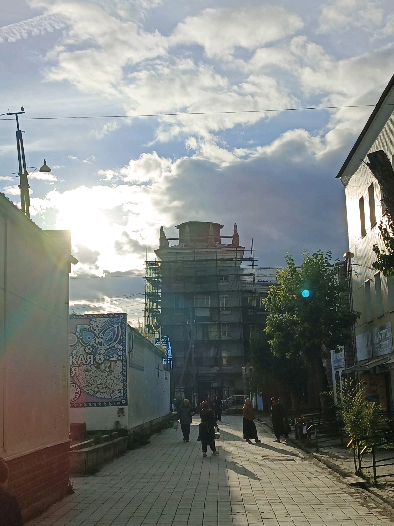 A sunny, cloudy day in Zlatoust, Russia. The image captures a street scene with several buildings, including a multi-story apartment building undergoing renovation, covered in scaffolding. The street is paved with interlocking bricks, and there are several people walking along the sidewalk. On the left side of the street, there is a white building with a mural on its side. On the right side, there are shops and trees. The sun is bright, casting long shadows on the ground. The overall impression is one of a busy urban environment on a pleasant day.