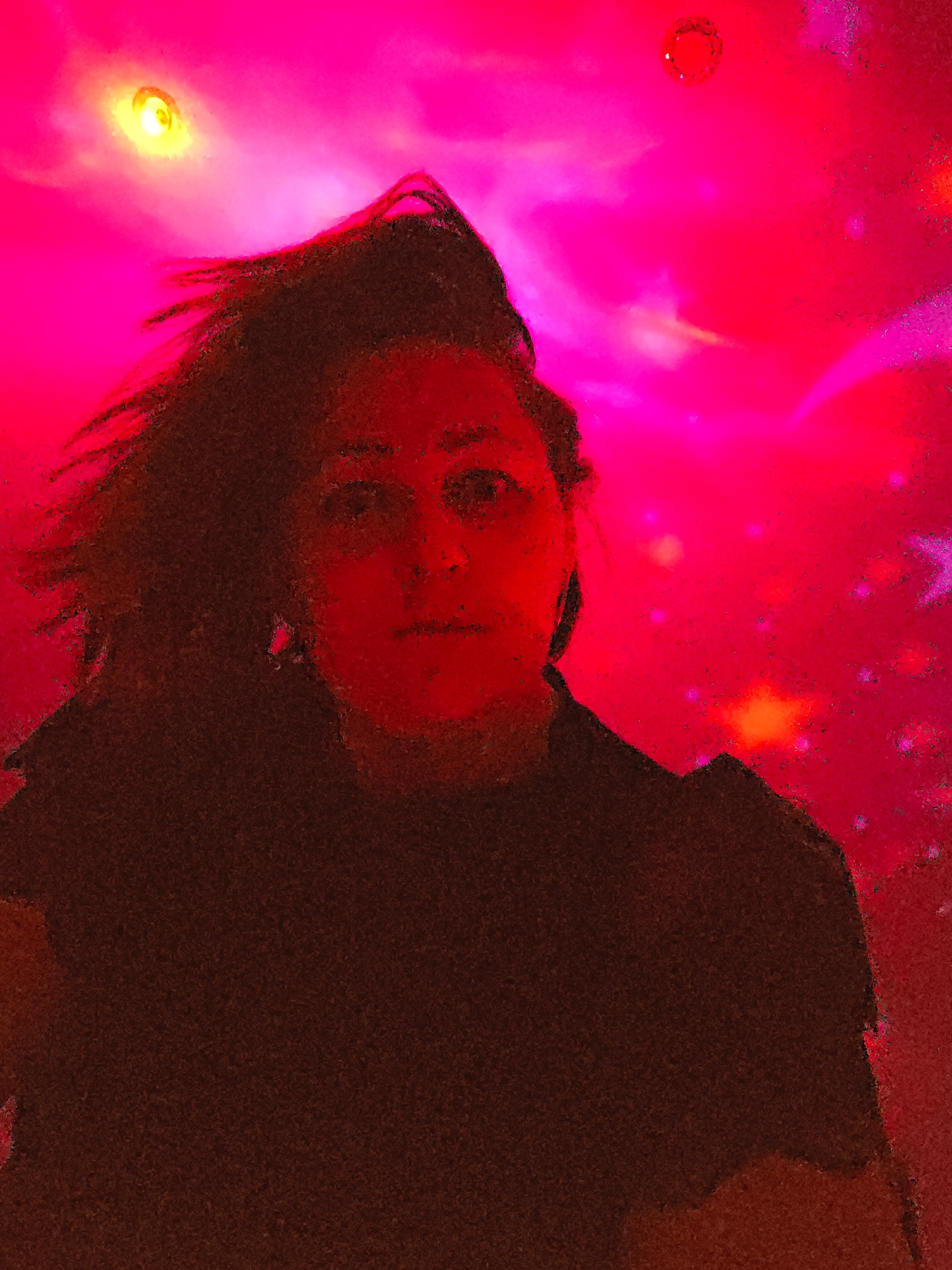 A person with dark hair and a dark shirt is facing the viewer. The background is a saturated mix of pink and red, with blurry, bright spots suggesting a light source. The image quality appears grainy. The location cannot be determined from the given image.