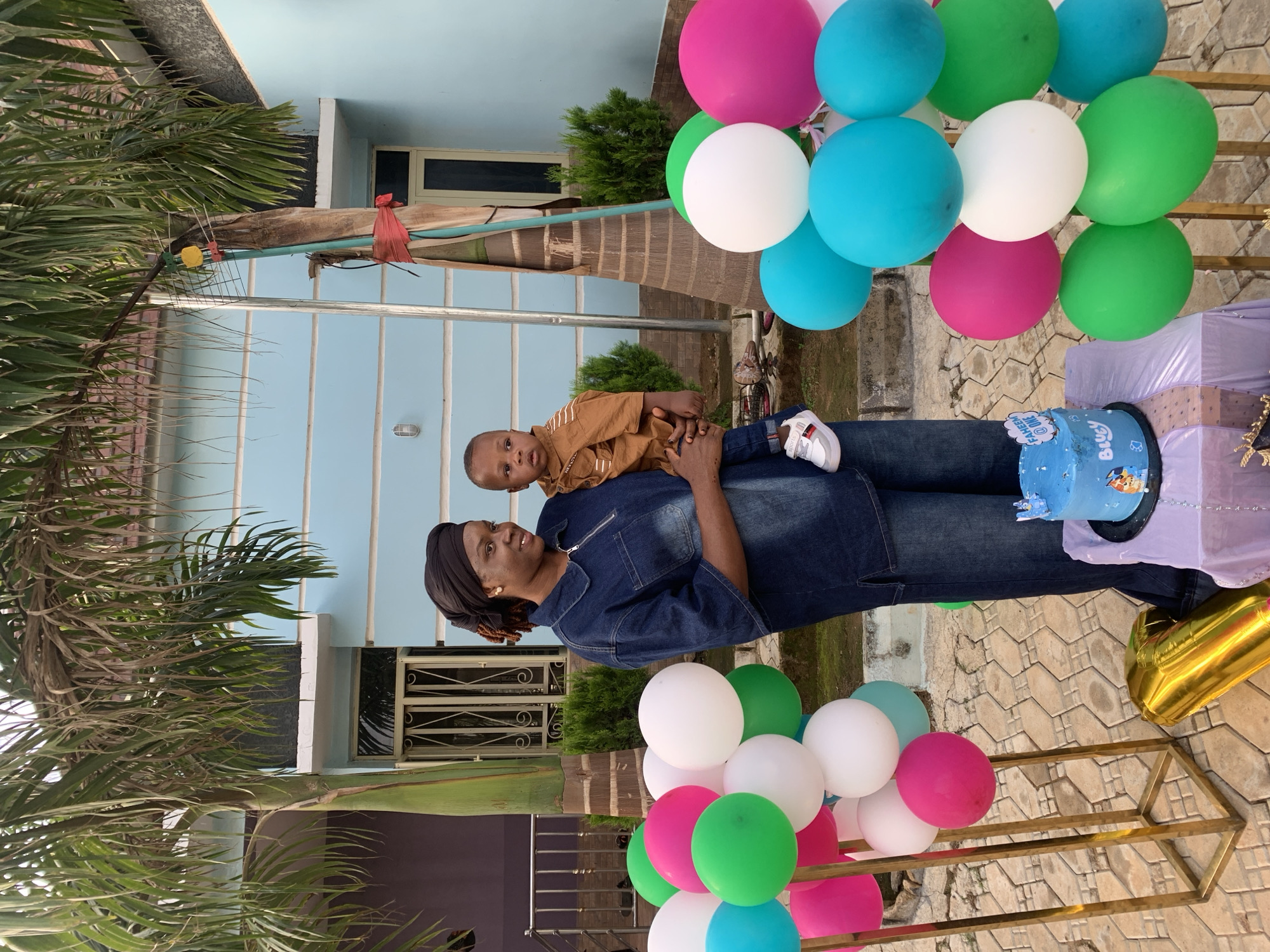 The image depicts an outdoor celebratory scene in what appears to be a residential area of Jos, Nigeria. The foreground shows a paved area constructed with interlocking brownish-grey tiles. A Black adult woman, likely in her late 20s or early 30s, stands centrally, holding a Black male infant. The woman wears a dark blue denim collared shirt with a zippered neckline and matching loose-fitting denim trousers. Her hair is covered by a black headwrap, and she wears small gold earrings. She smiles gently, looking towards the viewer's left. The infant, appearing to be around one year old, is dressed in a light brown collared long-sleeved shirt, dark blue jeans, and white sneakers with red accents; he looks directly forward with a neutral expression.
On either side of the woman and child are two gold-colored metallic vertical stands adorned with clusters of inflated balloons in bright pink, white, green, and light blue. To the right, at the bottom, a deflated gold foil balloon shaped like the number '1' lies on the pavement. Adjacent to it, a small, square table covered with a light lavender tablecloth holds a round, light blue cake decorated with characters resembling those from "Bluey" and topped with a white plaque reading "FAHEEM @ ONE."
In the background, a light blue building with white trim and security-barred windows is visible, partially obscured by two large palm trees. A darker, possibly purple, building is partially visible to the far left. Green shrubbery is present around the buildings and the bases of the palm trees. The palm tree on the right has a light blue pipe, a small metal grid with a yellow disc, and a piece of red fabric tied around its trunk. A bicycle is partially visible behind the left balloon stand. An outdoor wall-mounted light fixture is visible on the left side of the blue building.