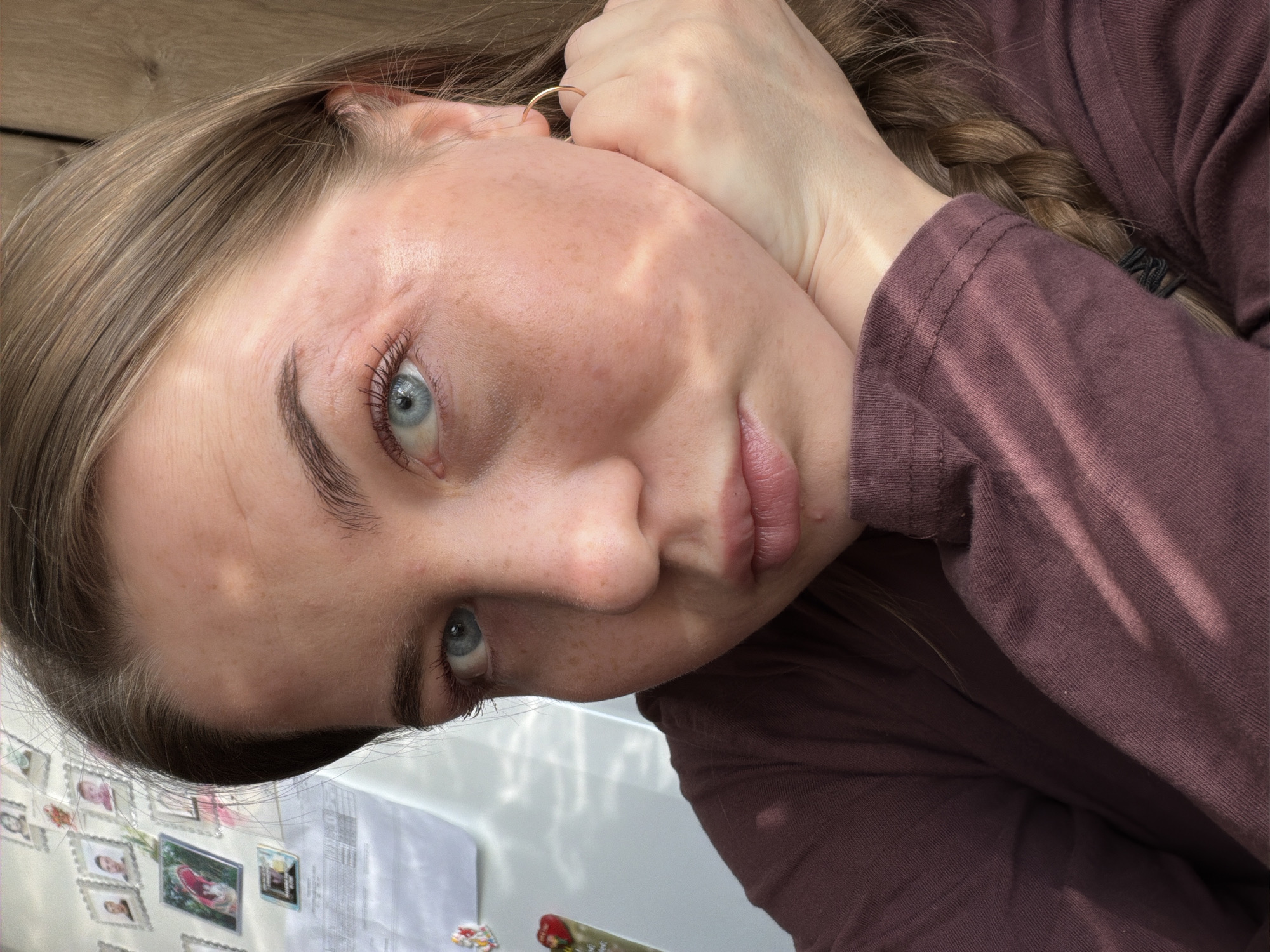 A close-up, angled selfie of a light-skinned female with freckles and blue eyes. Her light-brown hair is styled in a braid, and she is wearing a long-sleeved burgundy shirt. Her right hand is positioned near her face, and she is looking slightly downwards. The background includes a light-colored wooden surface and, partially visible, a white surface with various photos and documents affixed to it. The image does not contain enough information to place it in Уральское, Russia.