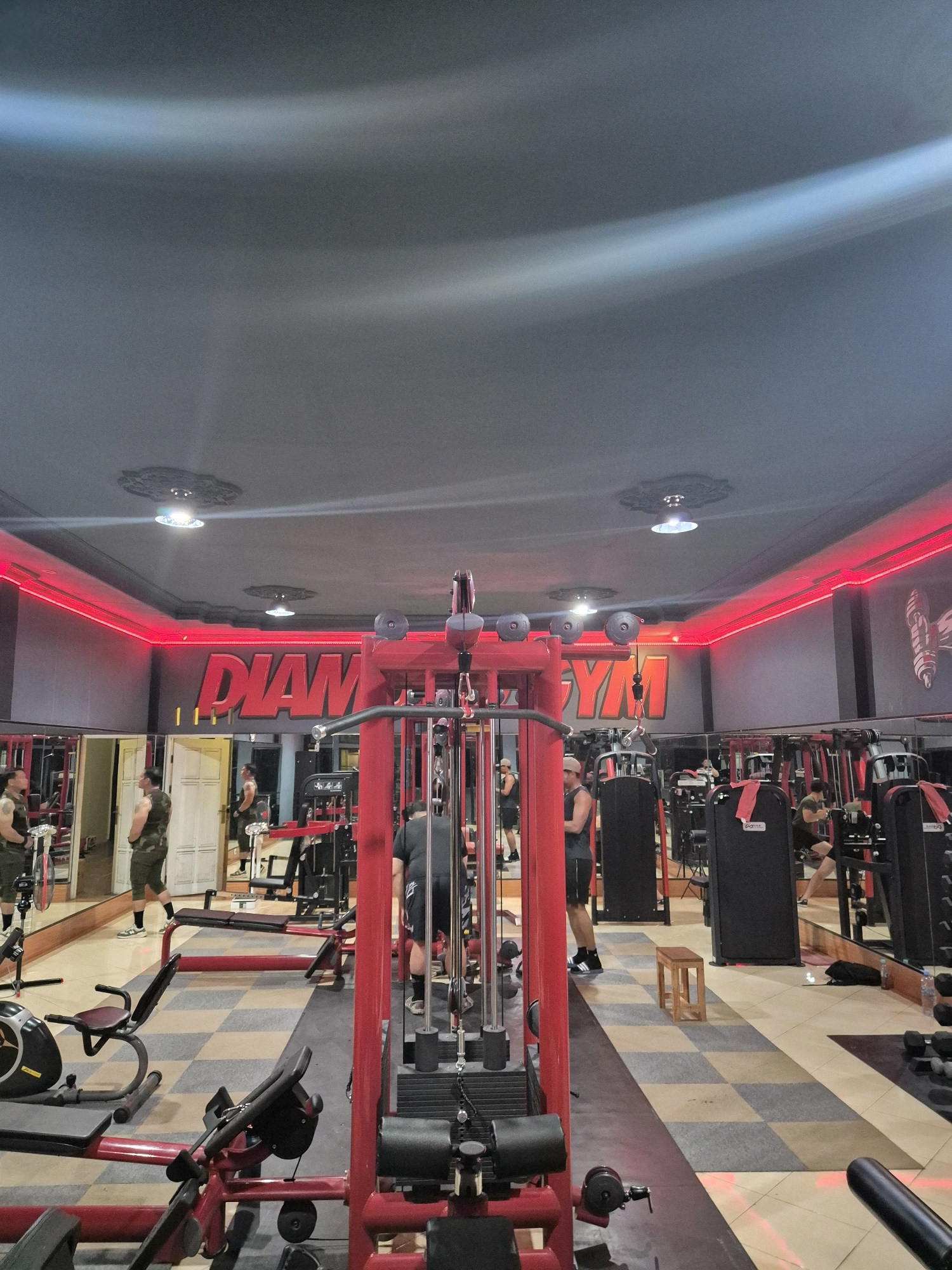 The image depicts the interior of a gym, identified as Diamond Gym, located in Bontang, Indonesia. The gym is equipped with various exercise machines, including weight machines, a rowing machine, and what appears to be a recumbent exercise bike. A prominent red sign with illuminated letters spelling "DIAMOND GYM" is mounted on the wall. Red LED lighting runs along the ceiling edges, illuminating the space. Several individuals are present in the gym, engaged in or preparing for workouts. One man is seen standing near a weight machine, another is walking, and a third is positioned in front of a mirror. Mirrors line the walls, reflecting the gym equipment and patrons. The floor is covered with checkered tiles in shades of tan and gray. The overall atmosphere suggests an active fitness facility.