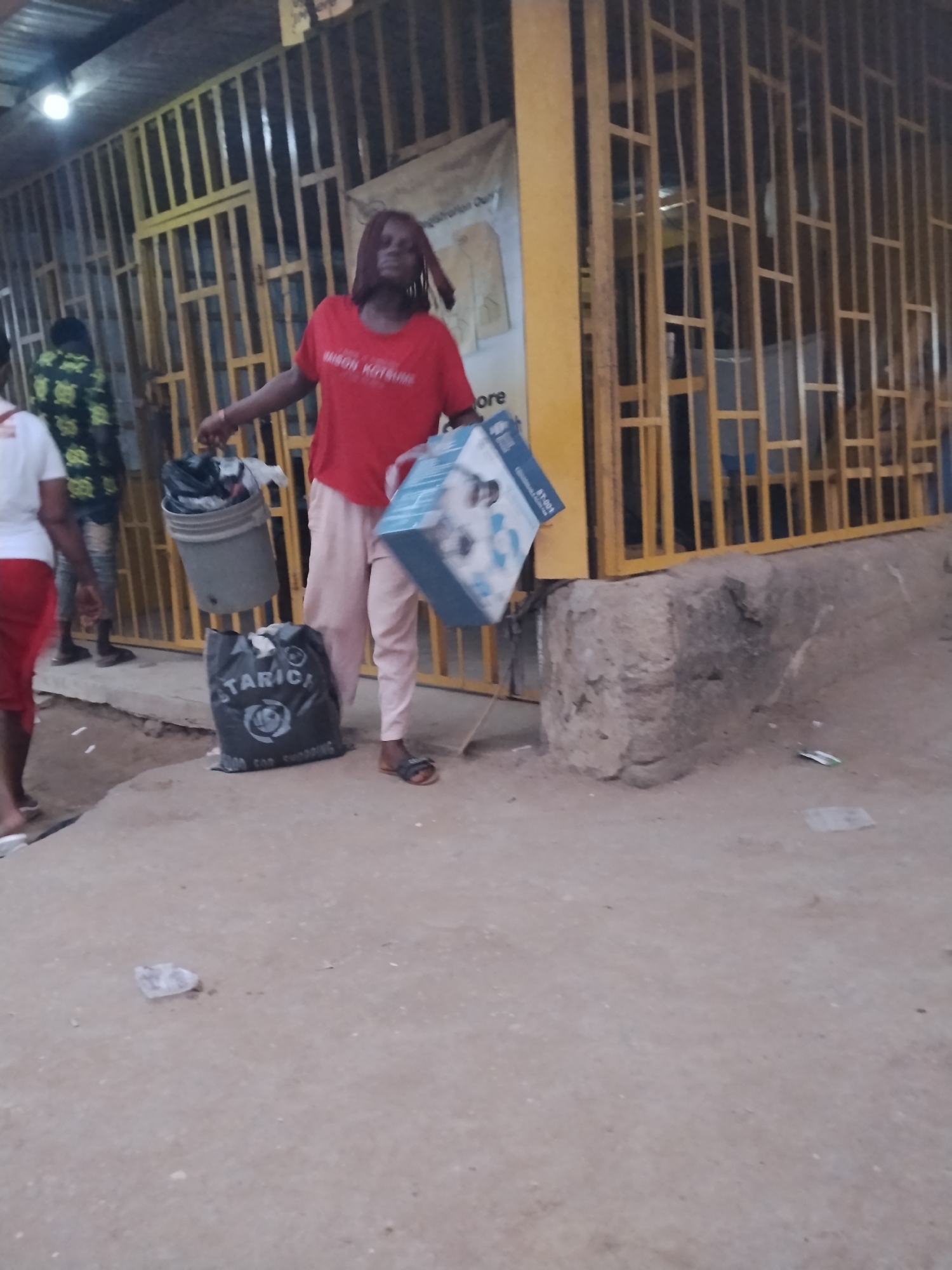 A woman stands in what appears to be a commercial area in Ura Nta Umuarandu, Nigeria. She is holding a grey bucket, a black shopping bag labeled "STARICH", and a blue packaged item. She is wearing a red t-shirt and light pink trousers. The ground is uneven and littered with debris. Behind her is a yellow storefront with a barred gate. Other people are vaguely visible in the background.