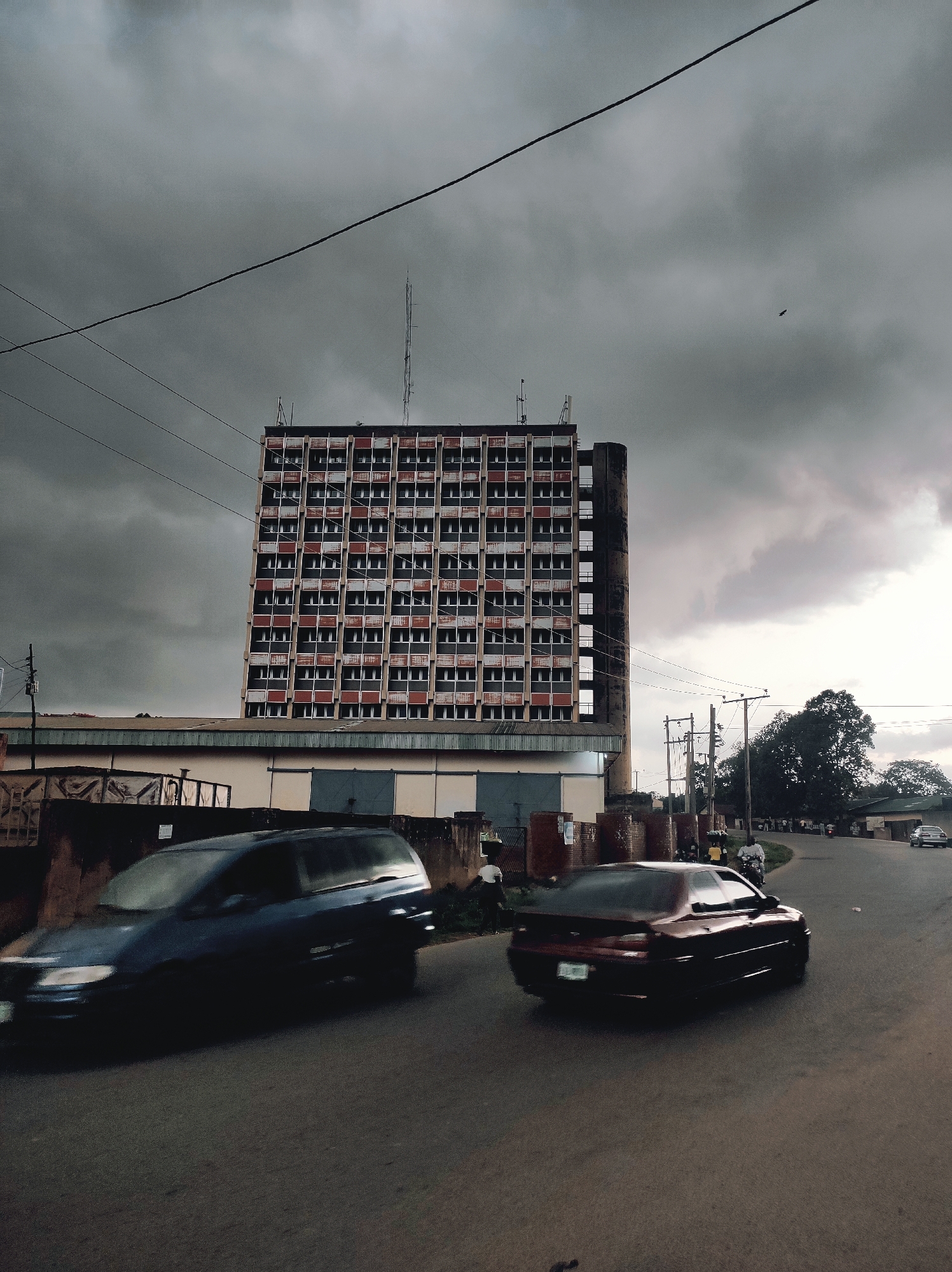 The image shows a street scene in Jos, Nigeria, dominated by a large, rectangular building with multiple rows of windows, likely an office or residential structure. The building is topped with a communication tower. In the foreground, two cars are moving on the road, a dark-colored van and a maroon sedan. The sky is overcast with dark, heavy clouds. Overhead power lines are visible. Additional people and vehicles can be seen further down the street. The overall lighting is dim and muted, suggesting a cloudy or overcast day.