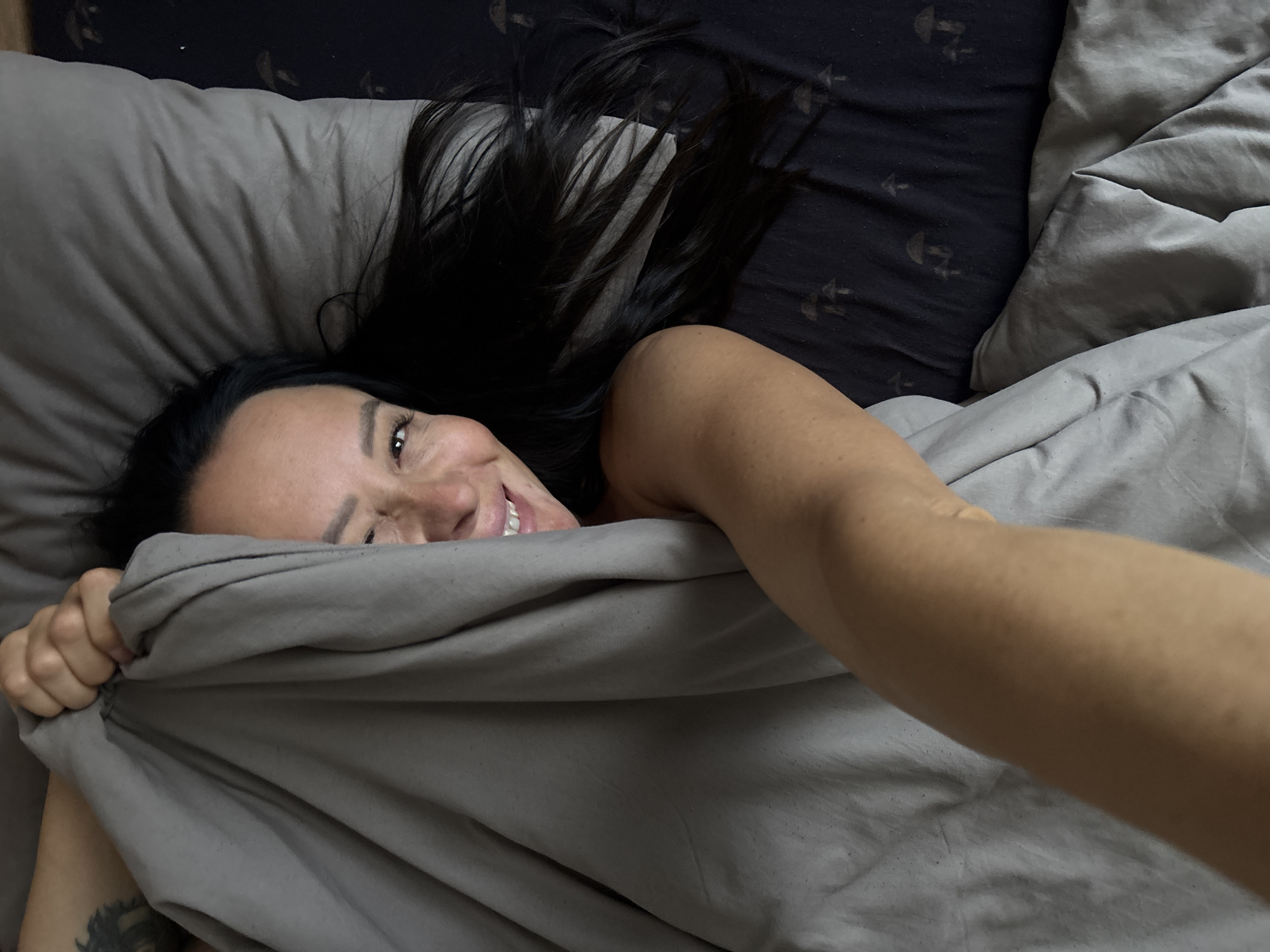 A woman is lying in bed, holding a grey blanket over her face. Only her eyes, nose, mouth, and part of her arm are visible. Her dark hair is spread out on a pillow. The bedding has a dark blue color with a pattern of mushrooms. The image appears to be taken from a high-angle perspective, looking down at the woman. There is no discernible location in Chaikovsky, Russia.