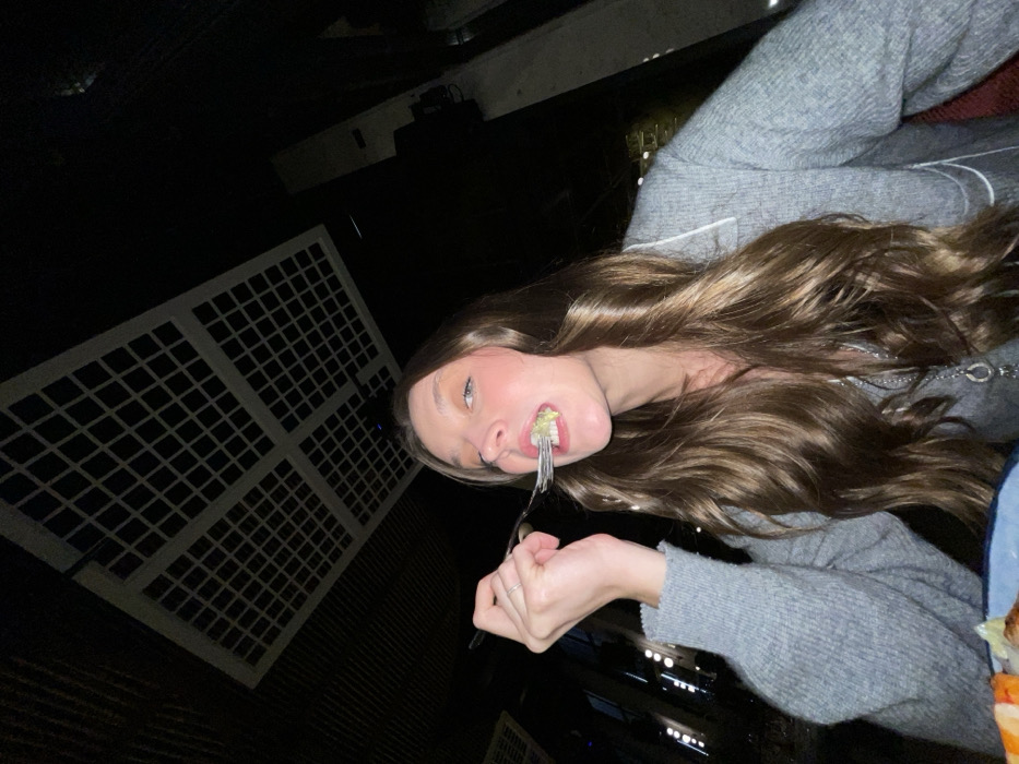 A young woman with long, light brown hair eats from a fork, tilting her head to the side. She wears a grey sweater. The background is dark, with a gridded, square pattern on the left side and faint hints of objects to the right. The scene has low lighting and appears to be outdoors at night.