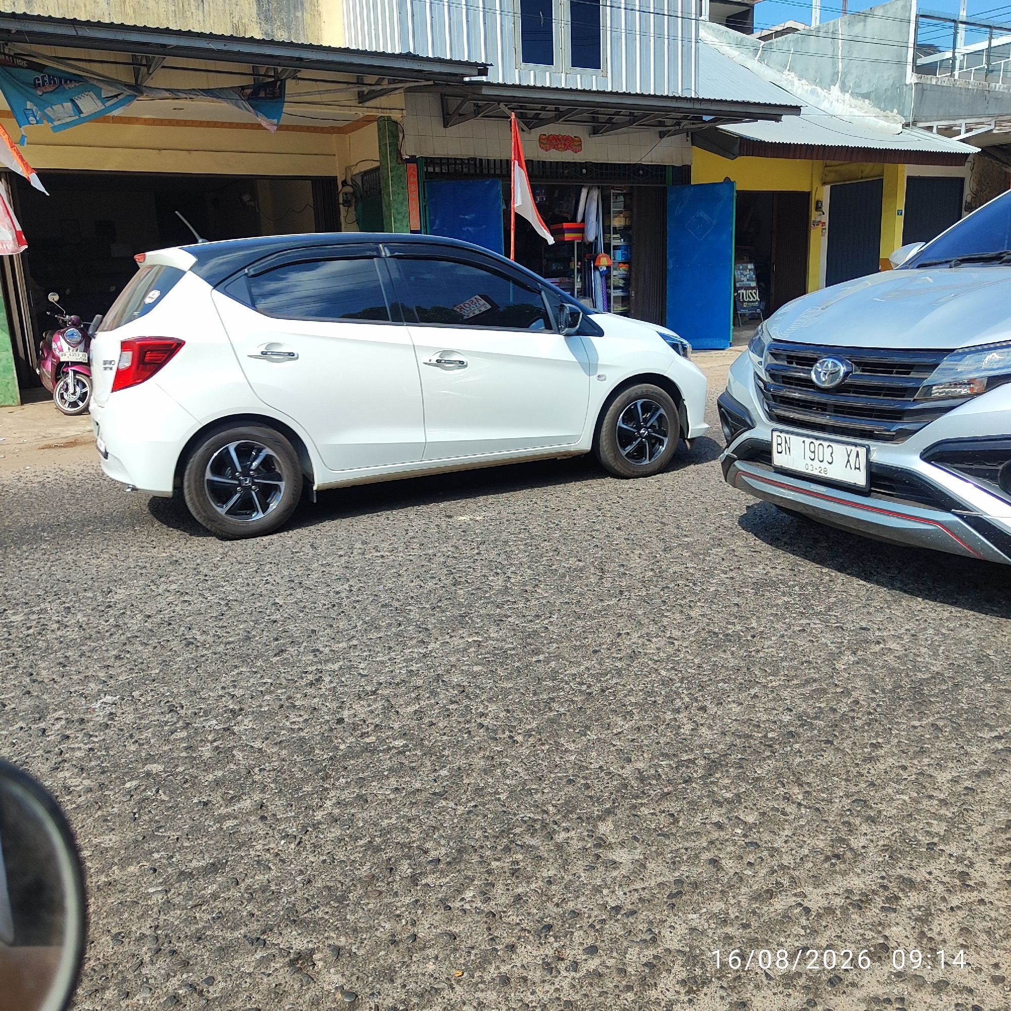 The image shows two cars parked on a paved road in front of several buildings. The building on the left appears to be a shop with an open doorway. A pink motorcycle is parked inside the doorway of the building on the left. The car on the left is white with a black roof, and the car on the right is silver. A license plate on the silver car reads "BN 1903 XA 03-28". There are flags and signs on the buildings, suggesting this is a commercial area. A timestamp in the bottom right corner indicates the date as 16/08/2026 and the time as 09:14. The scene is likely Pembaharuan, Indonesia, as stated in the prompt.