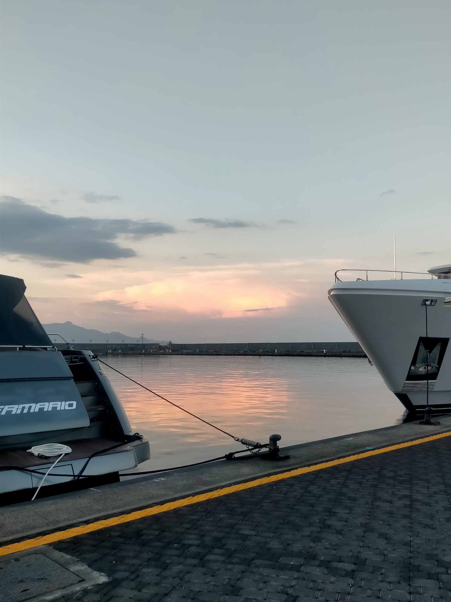 The image depicts a harbor scene at dusk, with a pale pink and orange sky visible behind a line of distant mountains and a breakwater. Two yachts are moored at a paved dock. The rear of a dark yacht, with the name "FERMARIO" visible, is in the foreground on the left. A white yacht's bow occupies the right side of the frame, angled away from the viewer. The water is calm, reflecting the colors of the sky. The dock is paved with dark rectangular stones and marked with a yellow line. Ropes and bollards are present for mooring. The scene is quiet and still, with no people visible.