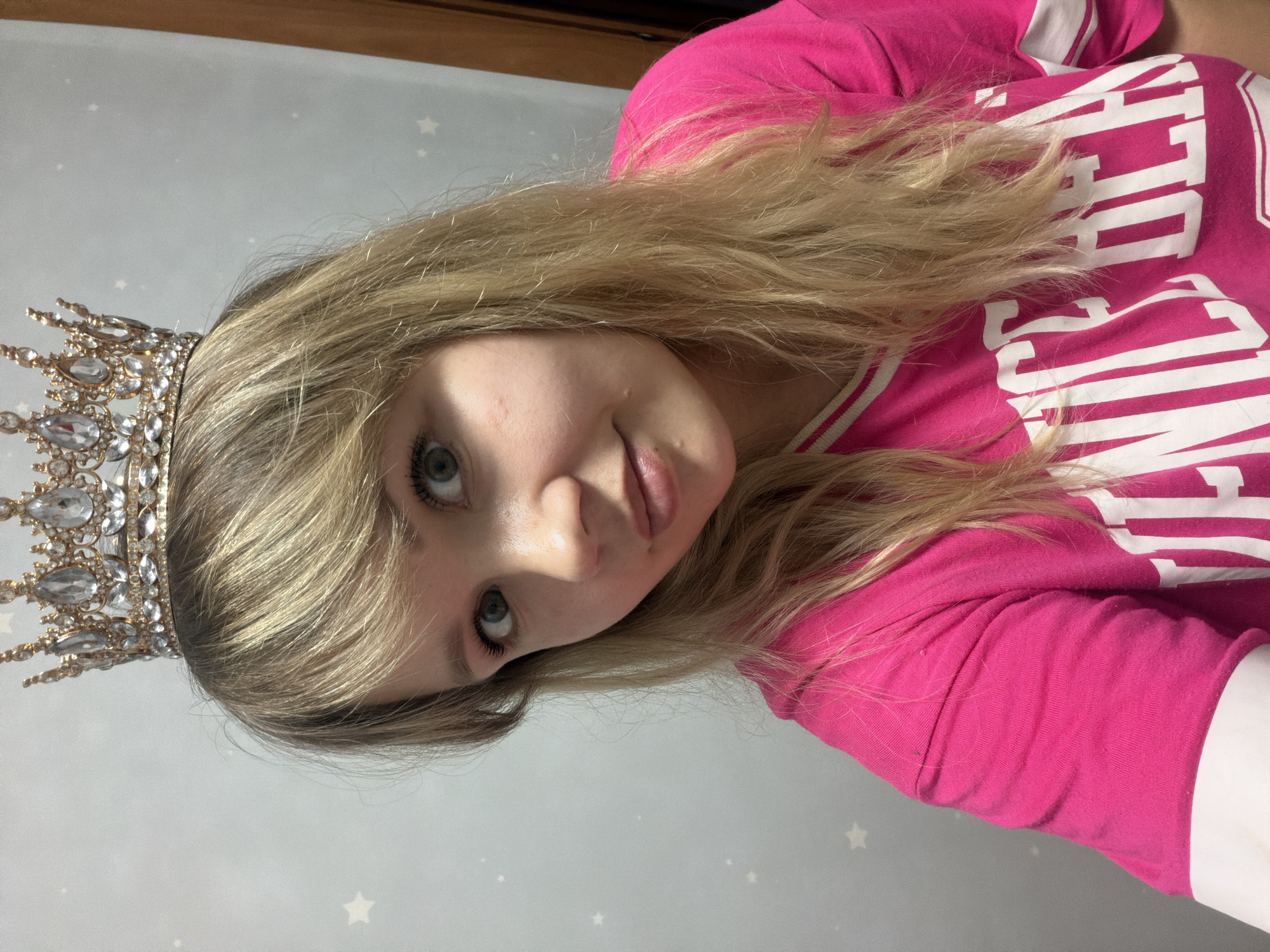 A young woman with light brown hair and green eyes, wearing a crown, is posing in a photograph. She is wearing a pink top with white lettering. The background features a light blue wall decorated with white stars. The location is Unknown, Unknown.