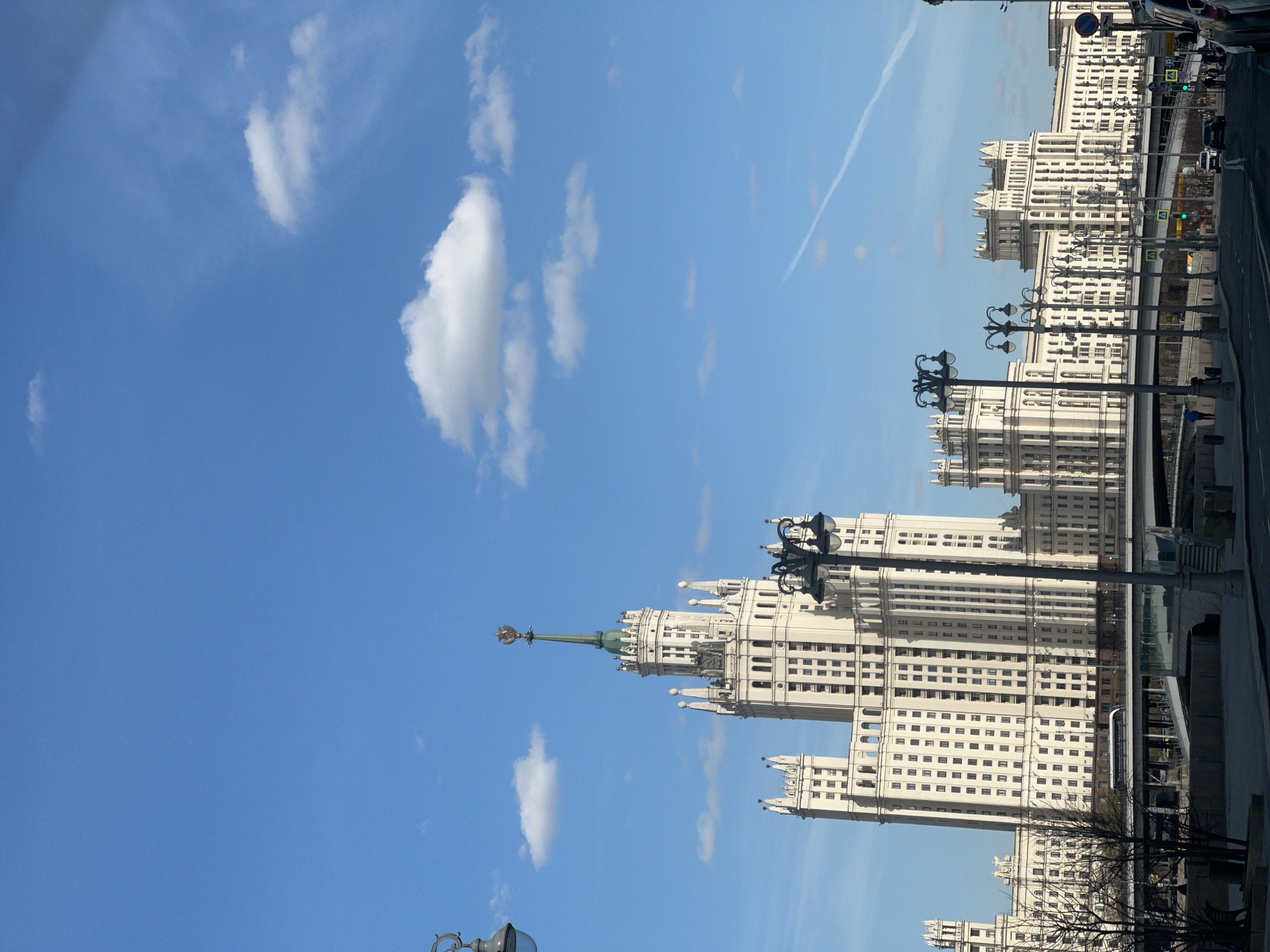 The image shows one of the Seven Sisters skyscrapers in Moscow, Russia, against a blue sky with scattered clouds. The Stalinist-era building is white with many windows and architectural embellishments, including a spire topped with a star or emblem. The building stands along a street with street lamps and other buildings in the background. There are also some cars and possibly pedestrians present, though they are not very detailed.