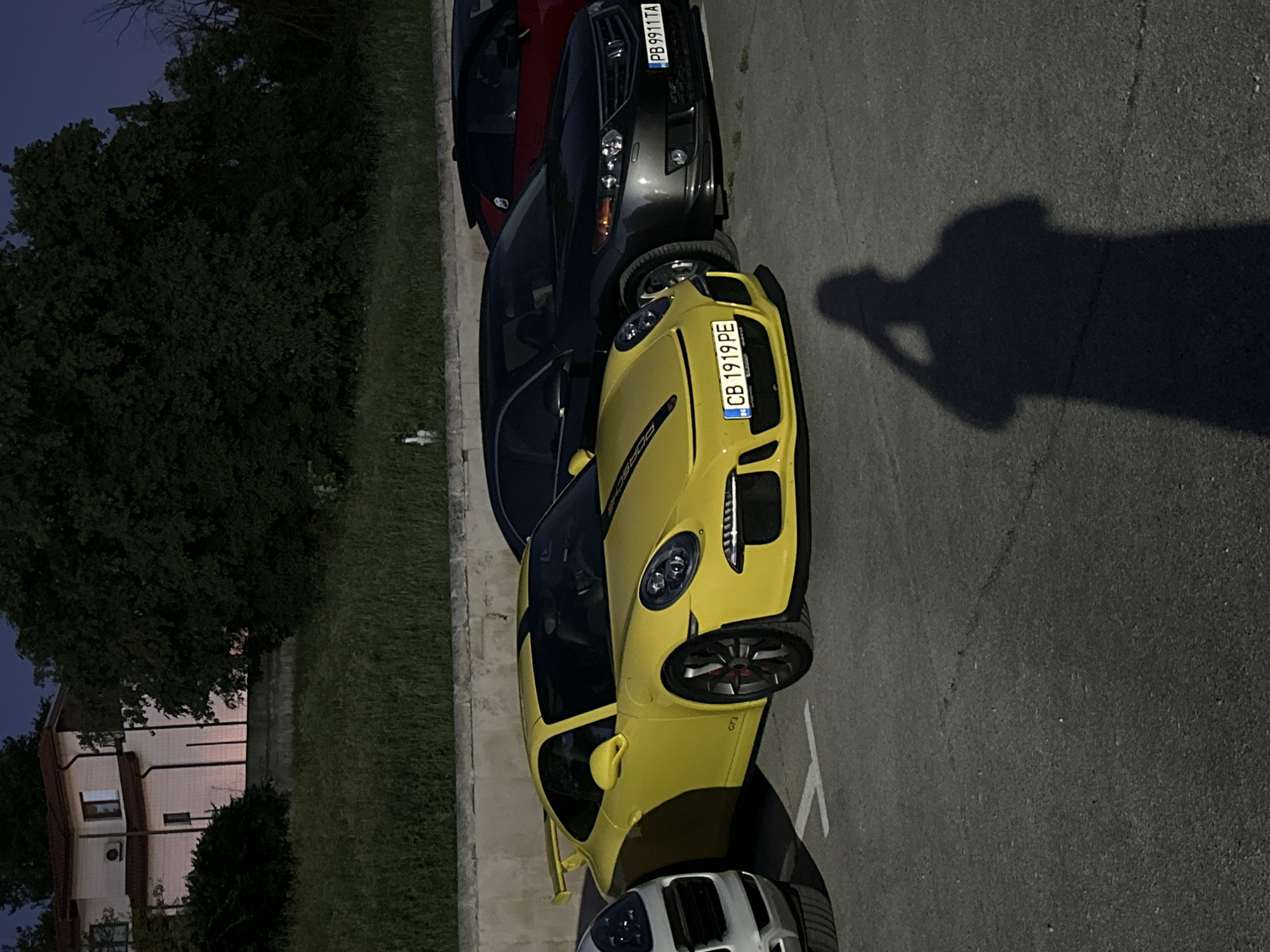 The image shows a yellow Porsche 911 GT3 parked in an asphalt parking lot. The car has black racing stripes on its hood and roof, and the license plate reads "CB 1919 PE". To the left of the Porsche is a white SUV, and behind it is a dark grey Honda Accord with the license plate "PB 9911 TA". Further back, a red car is partially visible. In the background, there is a building with a red roof and a lush green area with trees and grass. The scene appears to be in the city of Arbanassi, Bulgaria, as indicated by the building style and landscape. A shadow of a person holding a phone to take a selfie is cast on the asphalt in the foreground.
